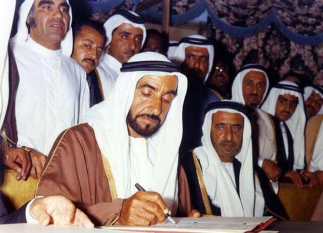 Sheikh Zayed bin Sultan Al Nahyan signs the historic Federation Agreement on December 2, 1971, officially creating the United Arab Emirates and ending the Trucial States era. On his left sits Sheikh Rashid bin Saeed Al Maktoum, Ruler of Dubai, with Mahdi Al Tajir, Sheikh Maktoum bin Rashid, and Sheikh Hamdan bin Rashid standing behind them. This momentous occasion marked the transition from British protectorate to independent nation.