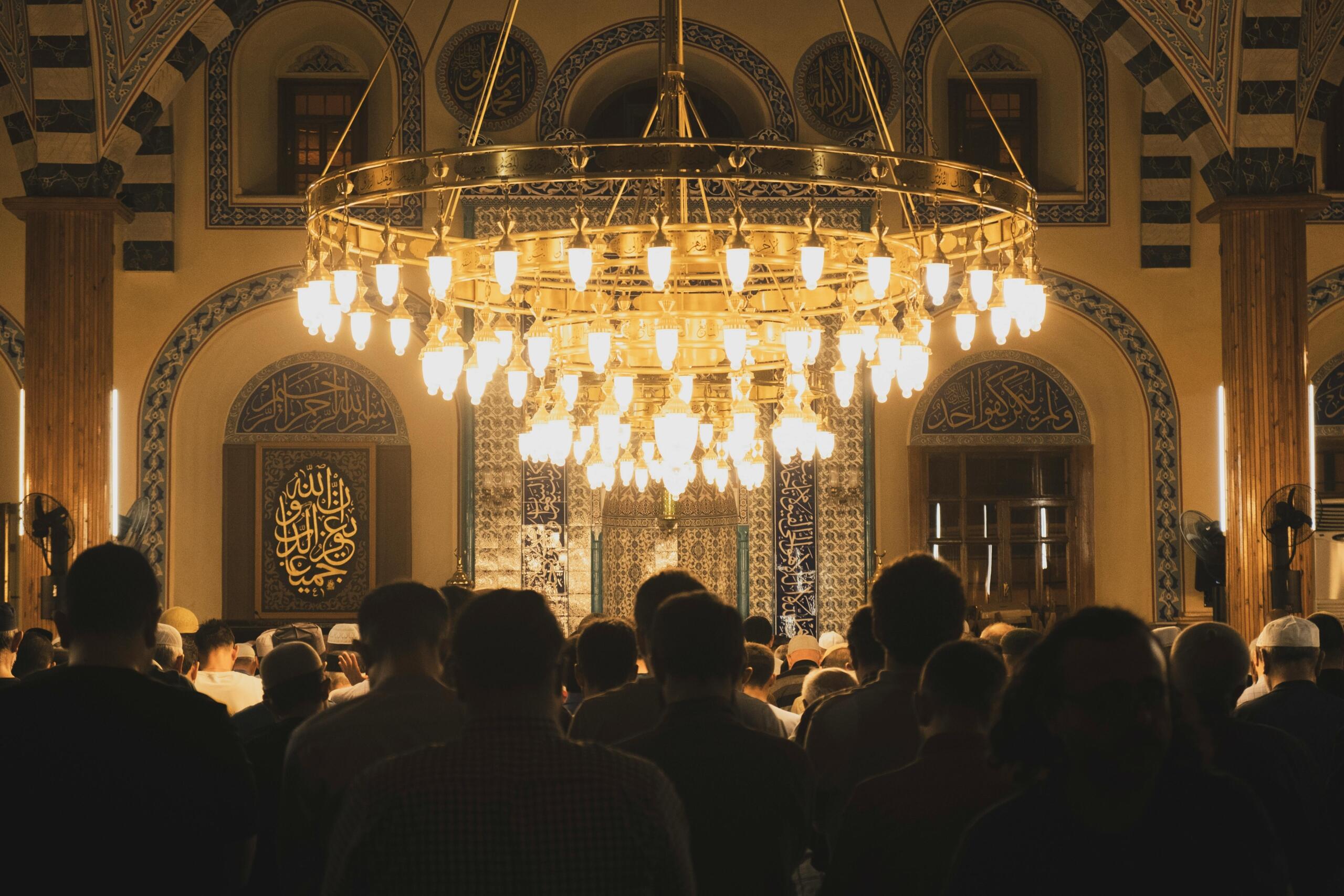 Muslims gathered in a mosque for prayer during the sacred Hijri months, with ornate chandelier and Arabic calligraphy