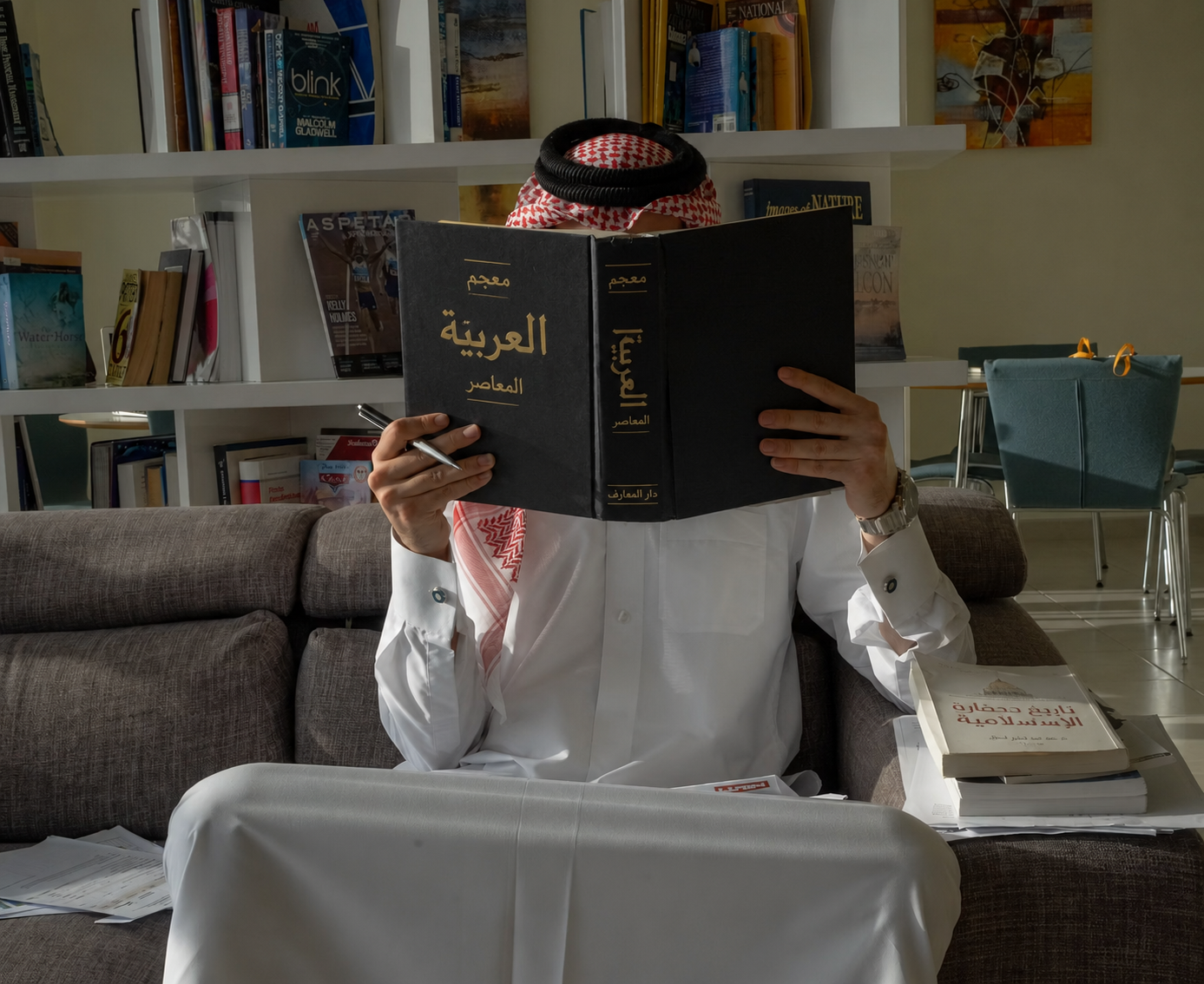 A man dressed in traditional Middle Eastern attire reads a modern Arabic dictionary in a contemporary library setting. This educational image represents language learning, academic research, literacy, Arabic culture, and personal development.