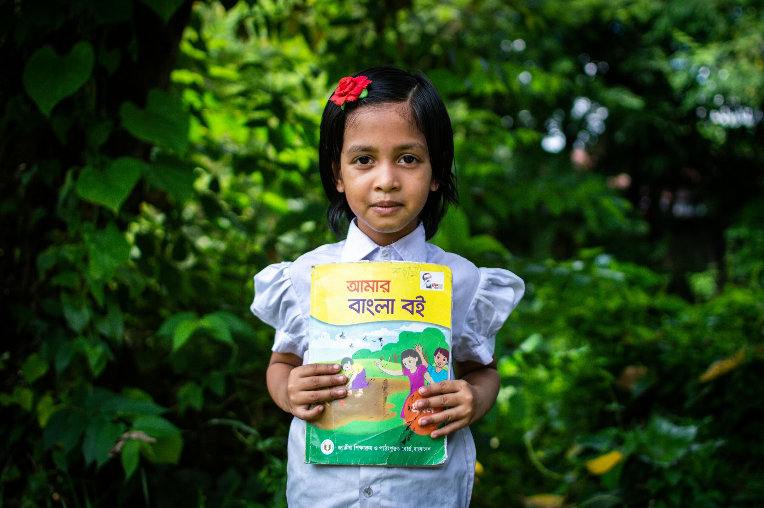 Bangladeshi girl in school uniform holding a Bengali language textbook titled Amar Bangla Boi