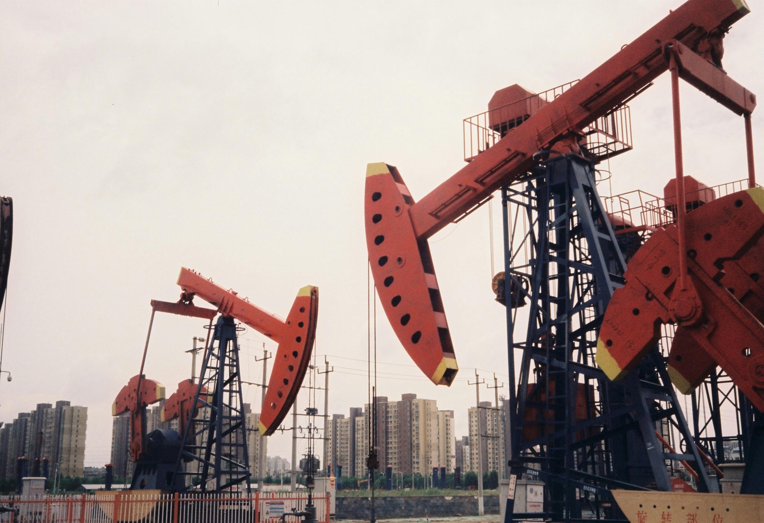 A close-up photograph of massive industrial pumpjacks with vibrant red arms and blue steel frames operating on an extraction field. In the background, a dense urban skyline with high-rise residential buildings stretches under an overcast sky.