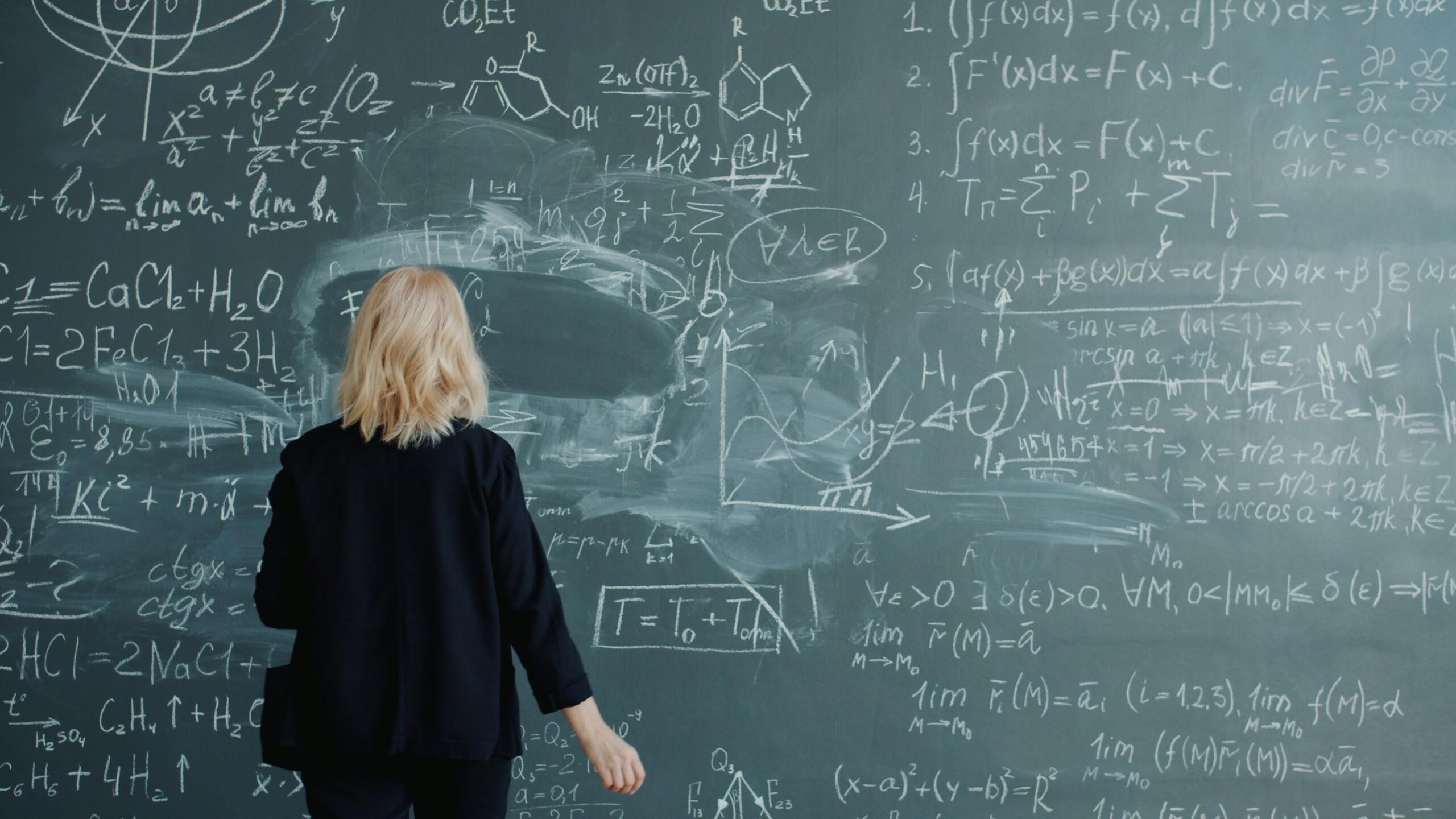 Person duke parë një tabelë të zezë me ekuacione të ndërlikuara matematike — momenti kur ia vlen të kërkosh ndihmë për algjebrën.