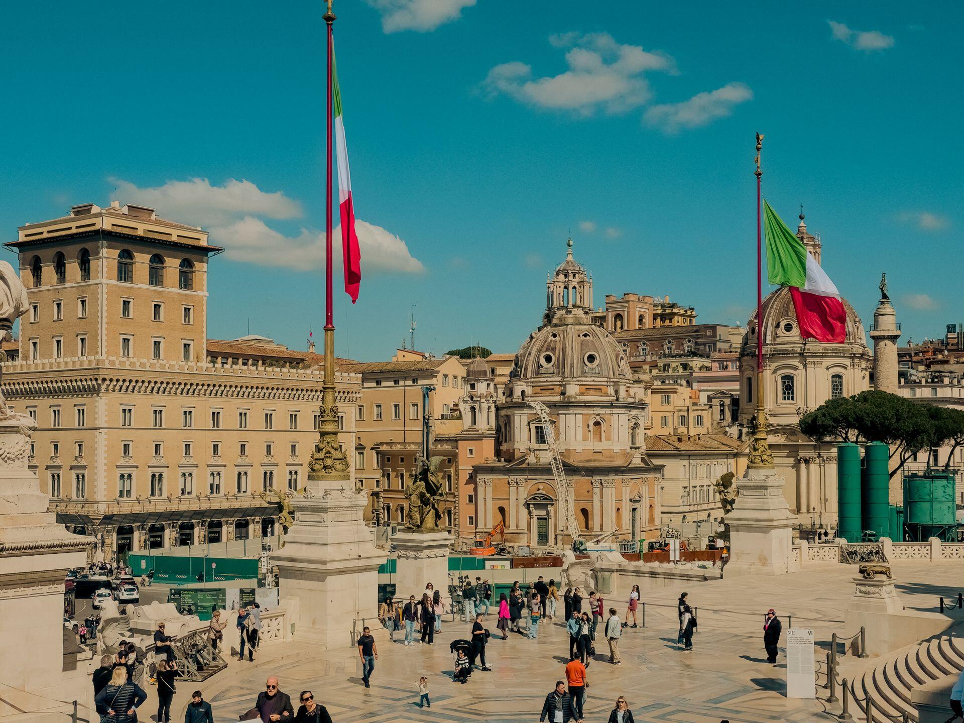 Vista de la Piazza Venezia en Roma, Italia, con banderas italianas flameando frente a edificios históricos y transeúntes. Fotógrafo: Paolo Bici.