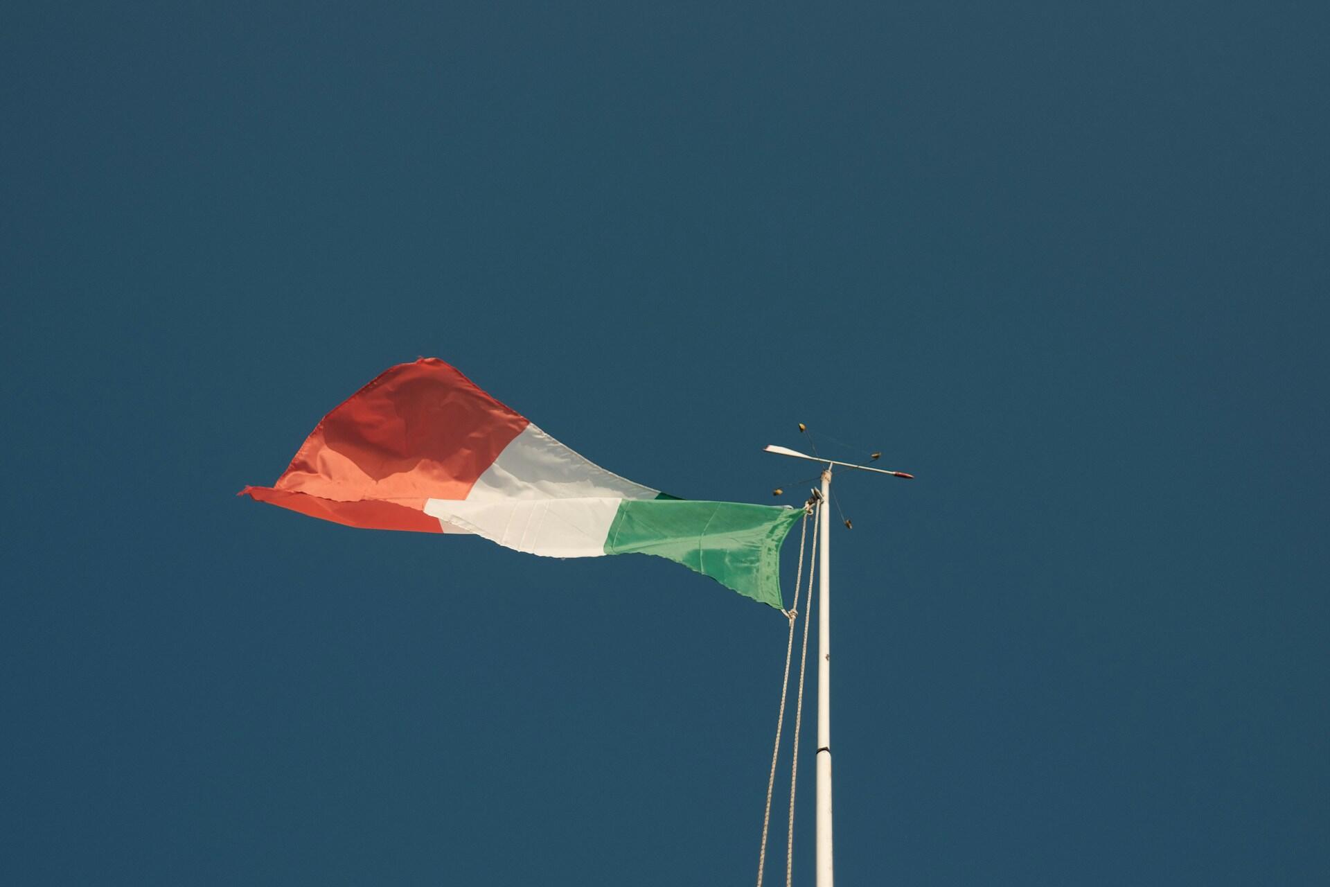 Primer plano de la bandera nacional de Italia ondeando en un mástil frente a un cielo azul despejado. Fuente: Unsplash.