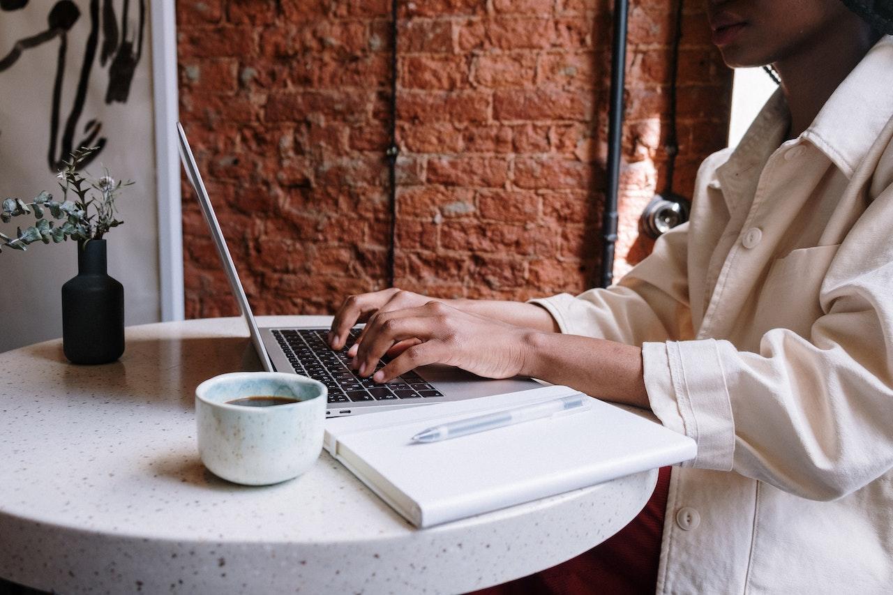 Mujer escribiendo en la computadora con una libreta y una taza de café. 