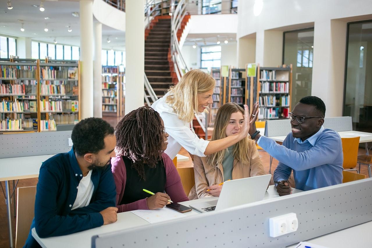 Alumnos en la biblioteca con sus computadoras, chocando las manos.