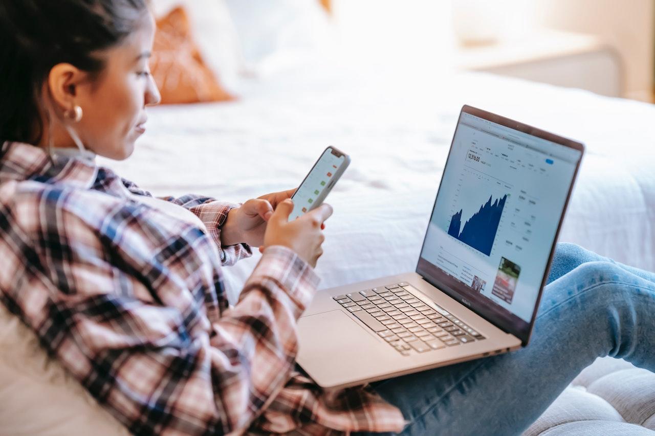 Mujer con la computadora abierta sobre sus piernas, y celular en la mano.