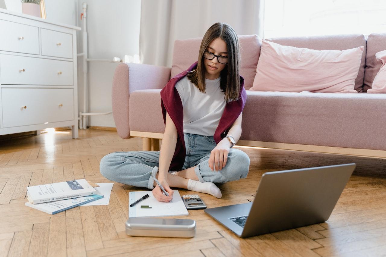 Joven estudiando sentada en el suelo con la computadora abierta y papeles.