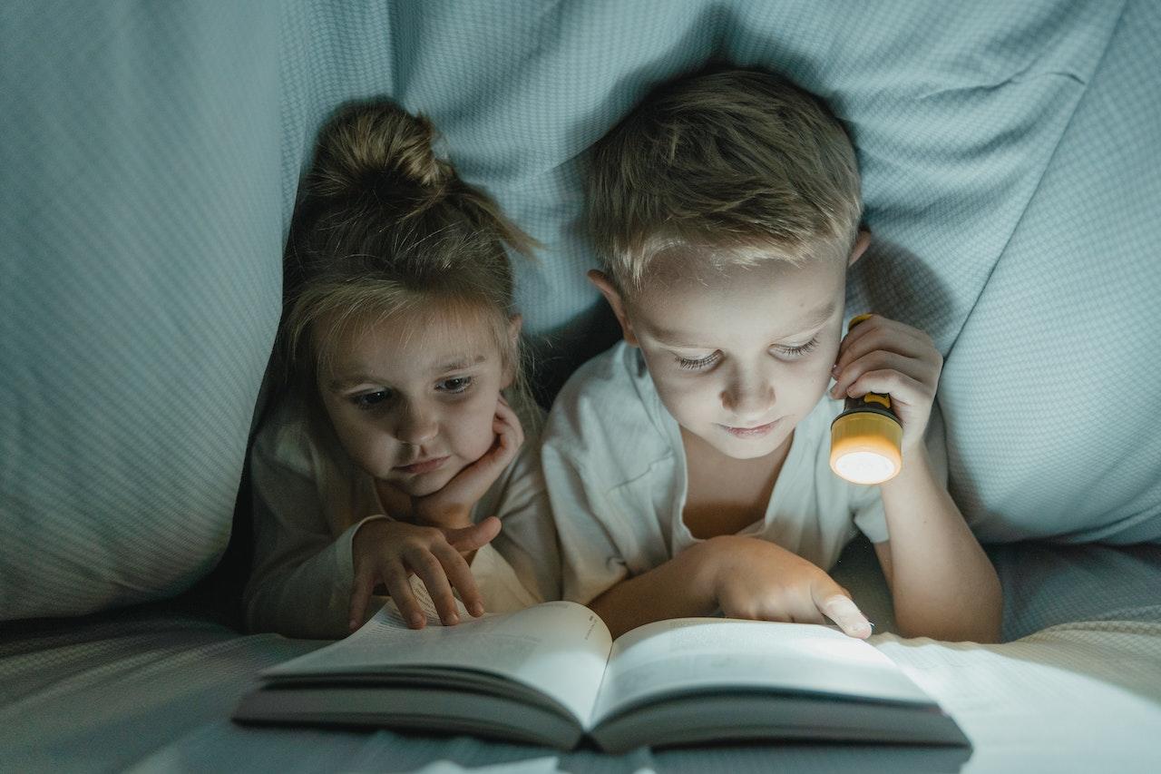 Niños leyendo un libro bajo una frazada, alumbrando con una linterna.