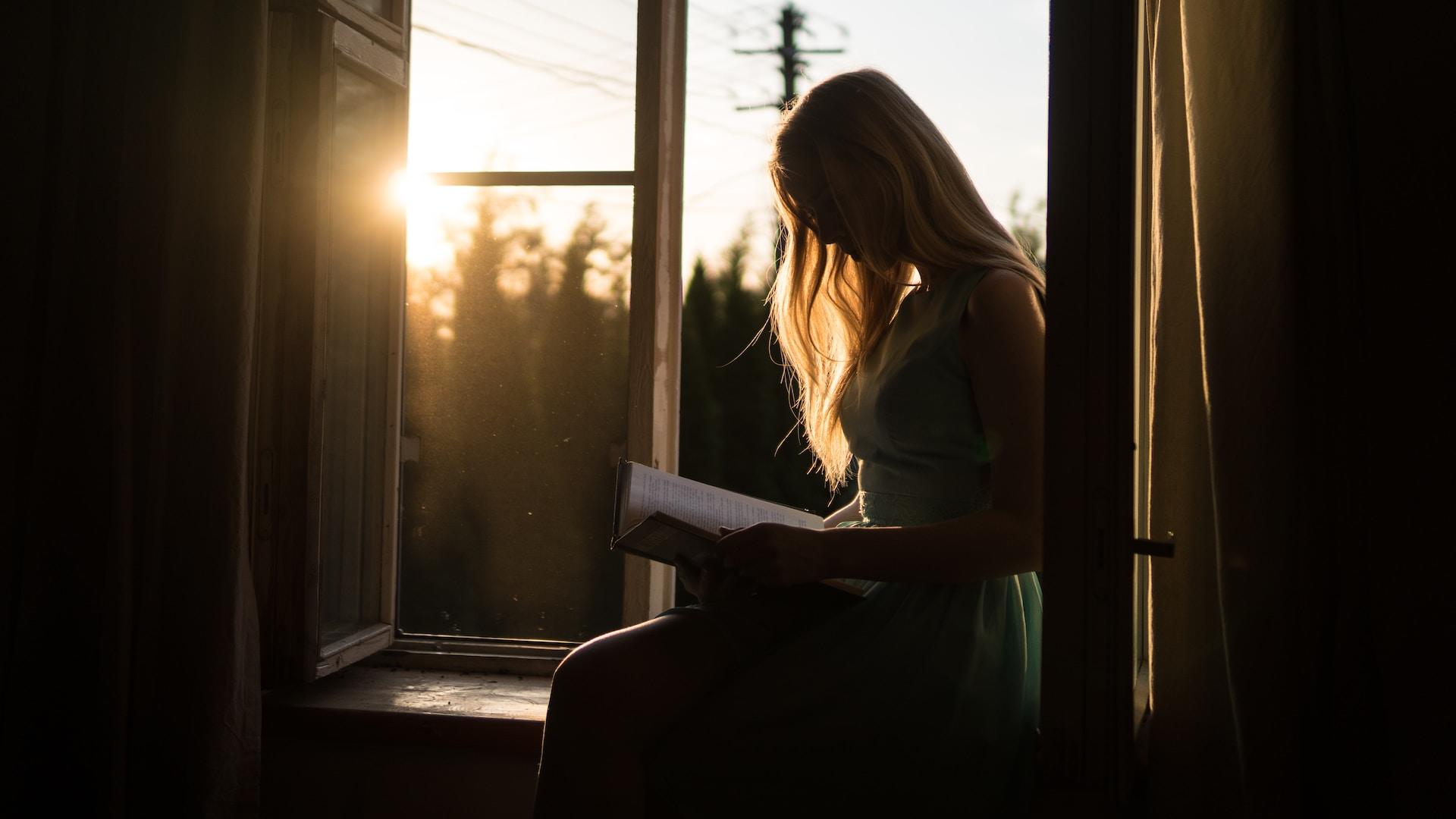 Mujer leyendo un libro sobre el marco de la ventana.