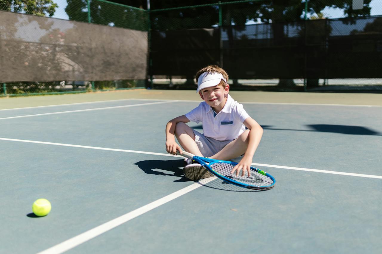 Niño vestido de blanco, sentado en una cancha de tenis con una raqueta en la mano.