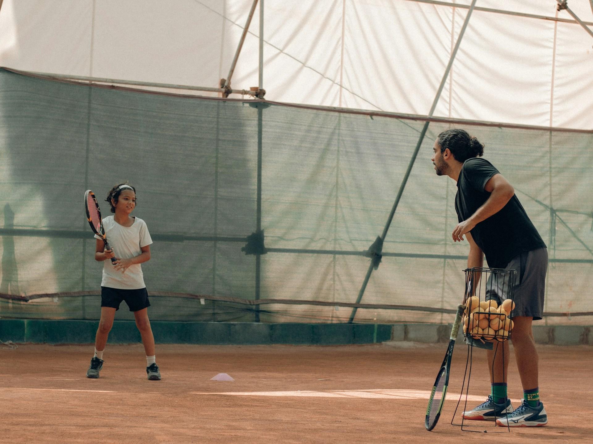 Hombre y niña en la cancha de tenis con raquetas y canasto de pelotas.