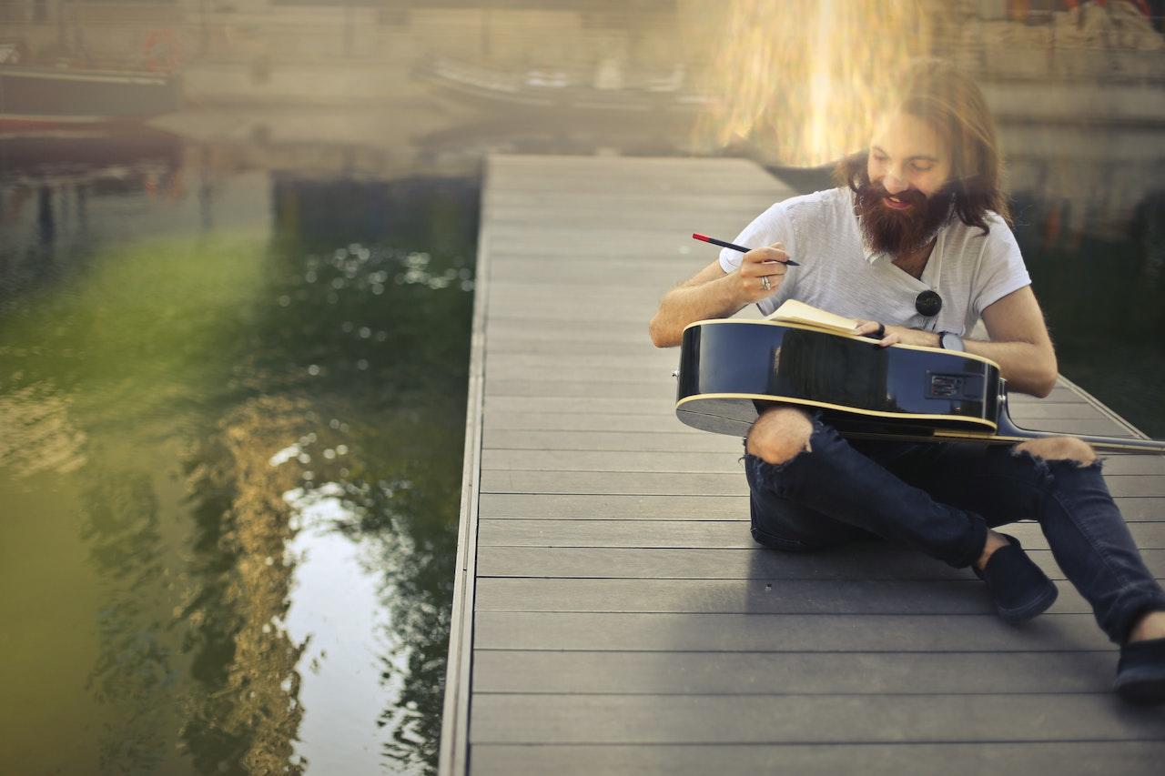 Hombre sentado en el puente de madera escribiendo sobre la guitarra.