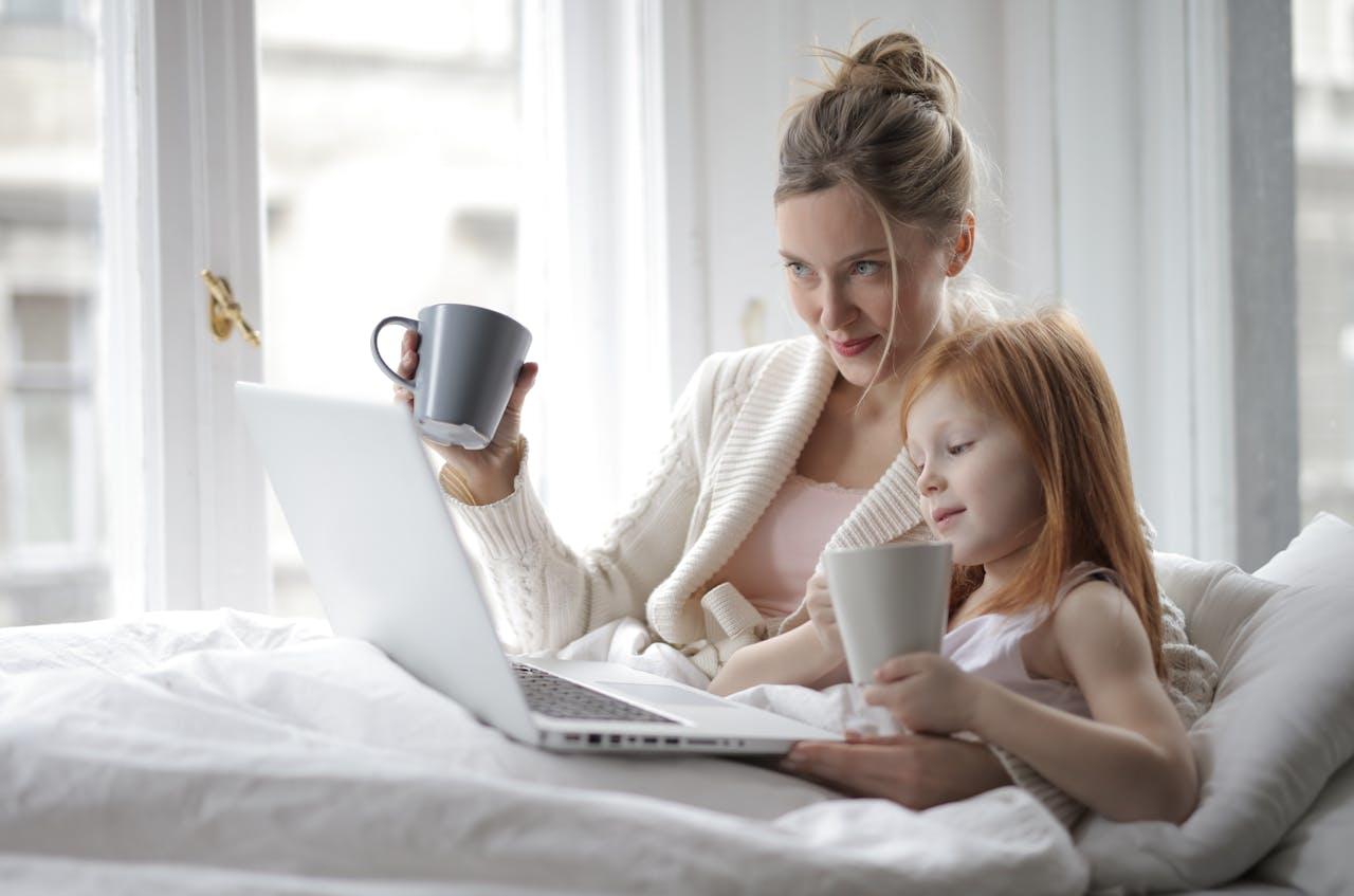 Mujer y niña en la cama con una taza en la mano, mirando la computadora.