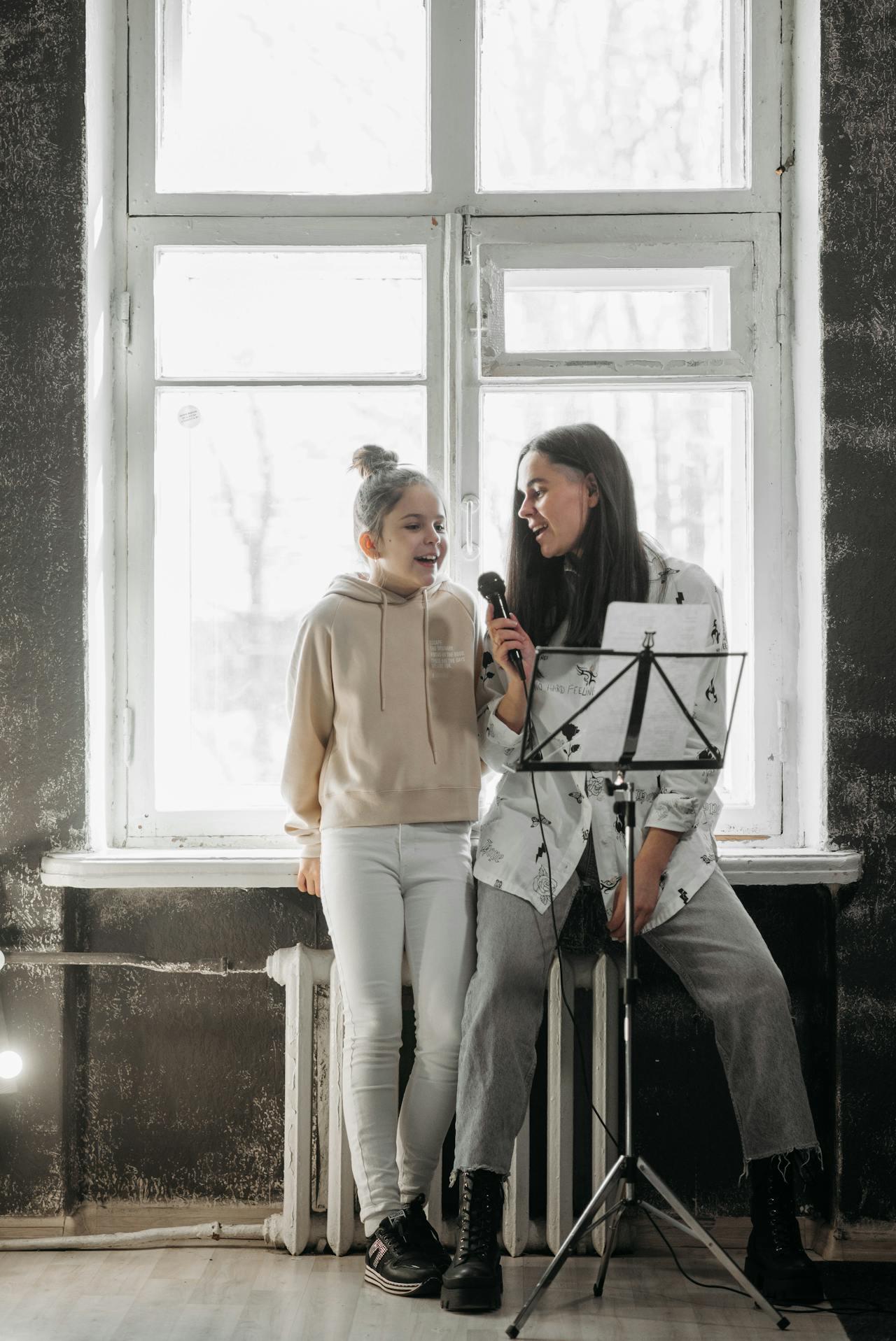 Niña y adulta cantando con micrófono paradas contra la ventana.