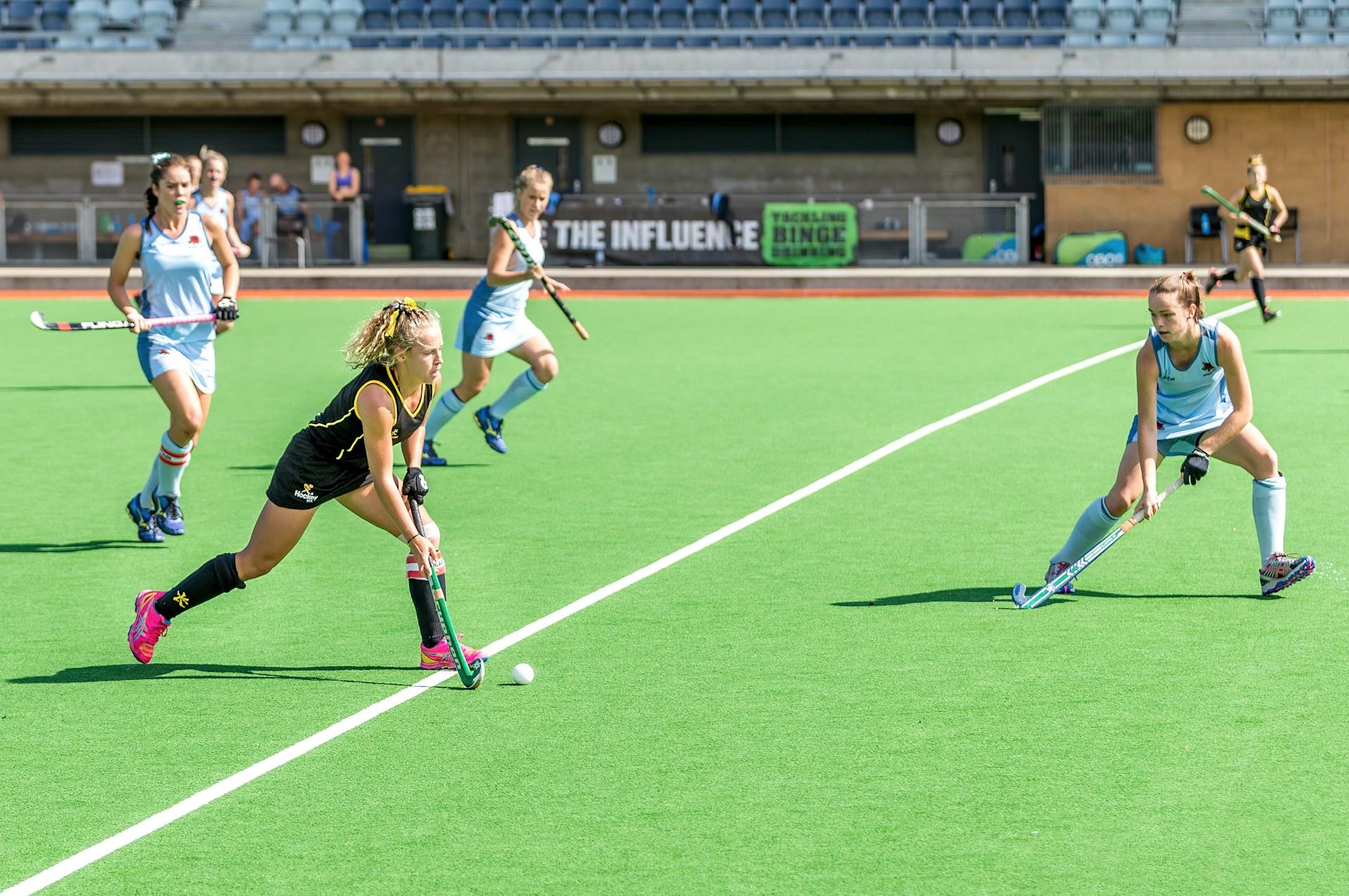 Mujeres con camisetas negras y celestes jugando en el campo con palos de hockey.