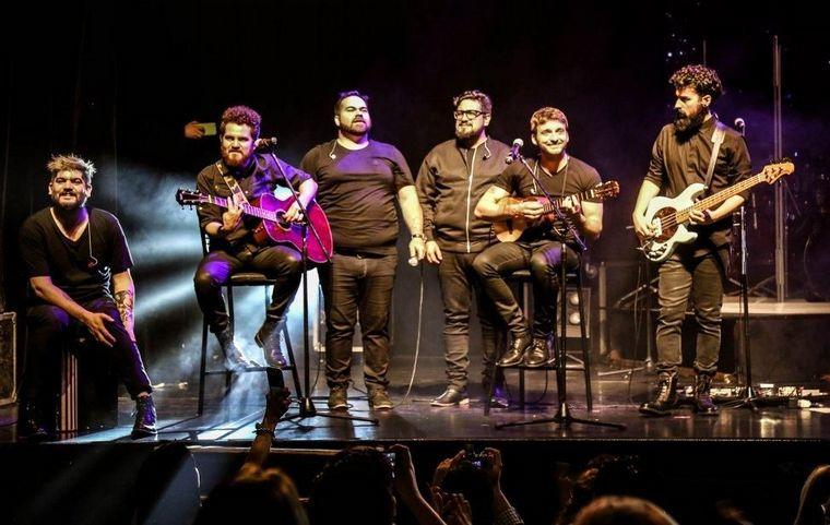 Seis hombres vestidos de negro con instrumentos, sobre el escenario.