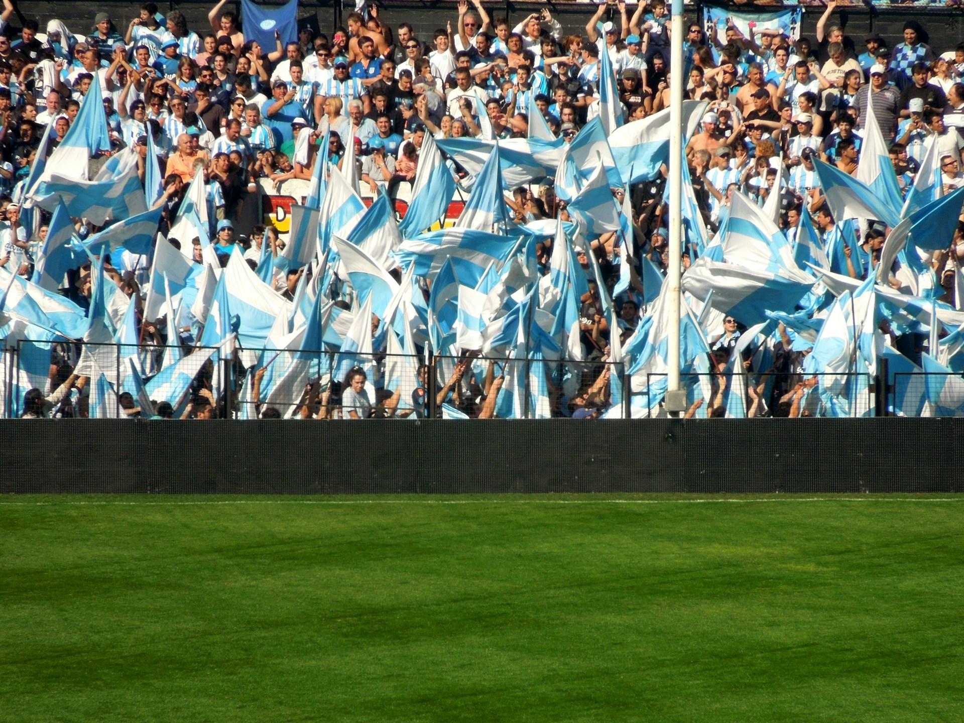 Hinchada en estadio con banderas celestes y blancas.