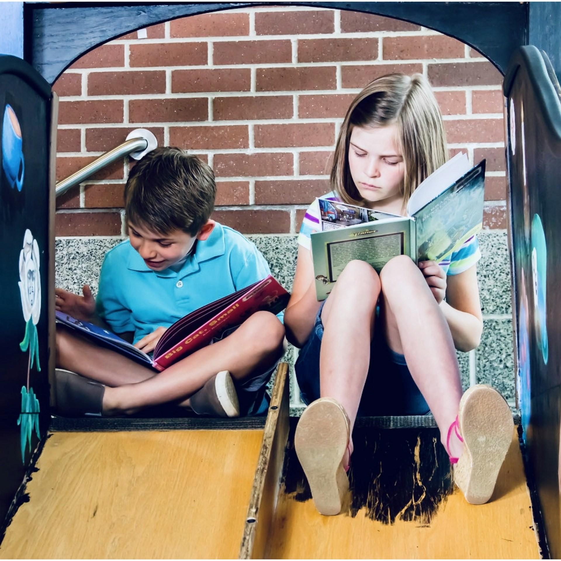 Una niña y un niño leyendo libros sentados en el tobogán.