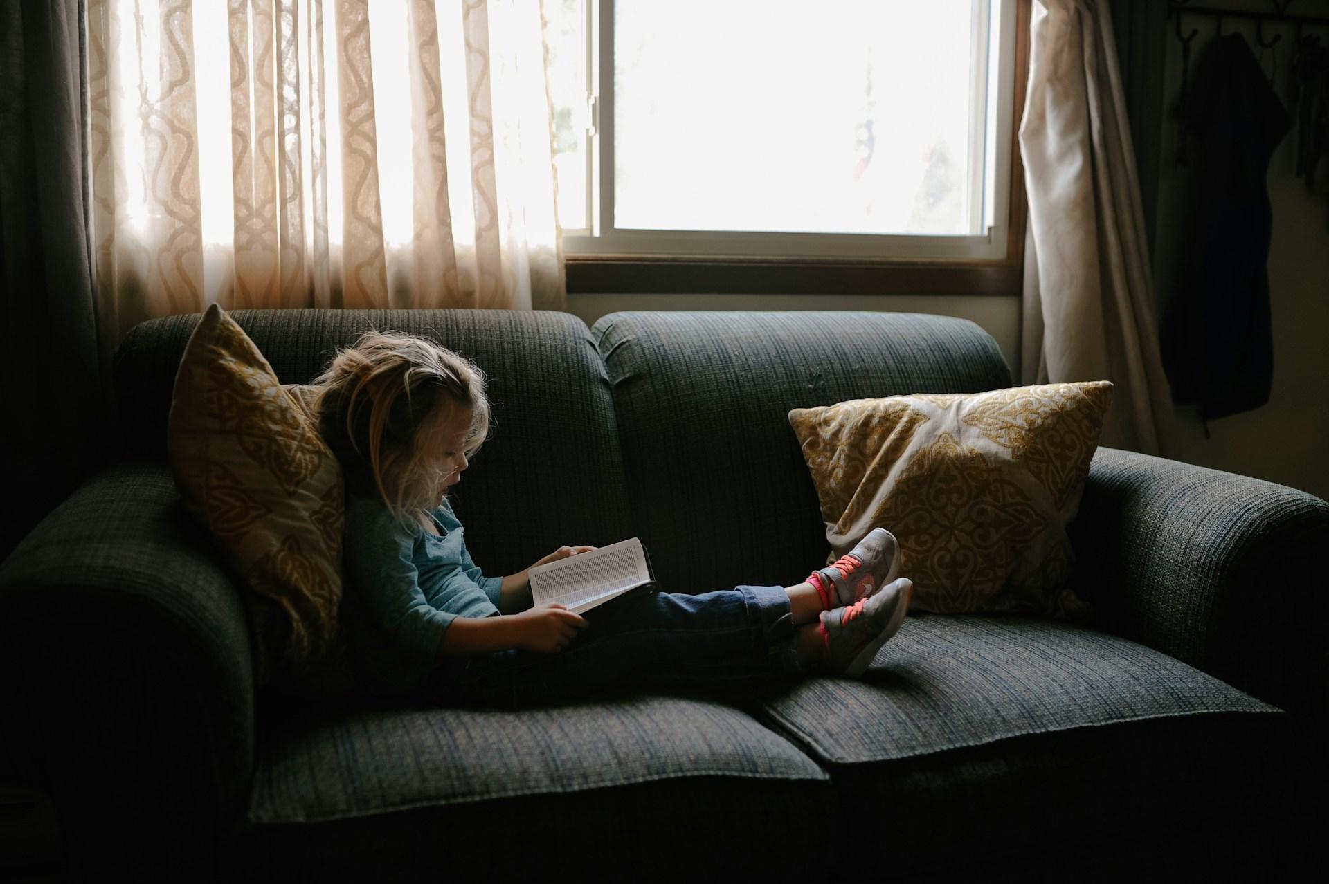 Niña leyendo un libro en el sillón gris.