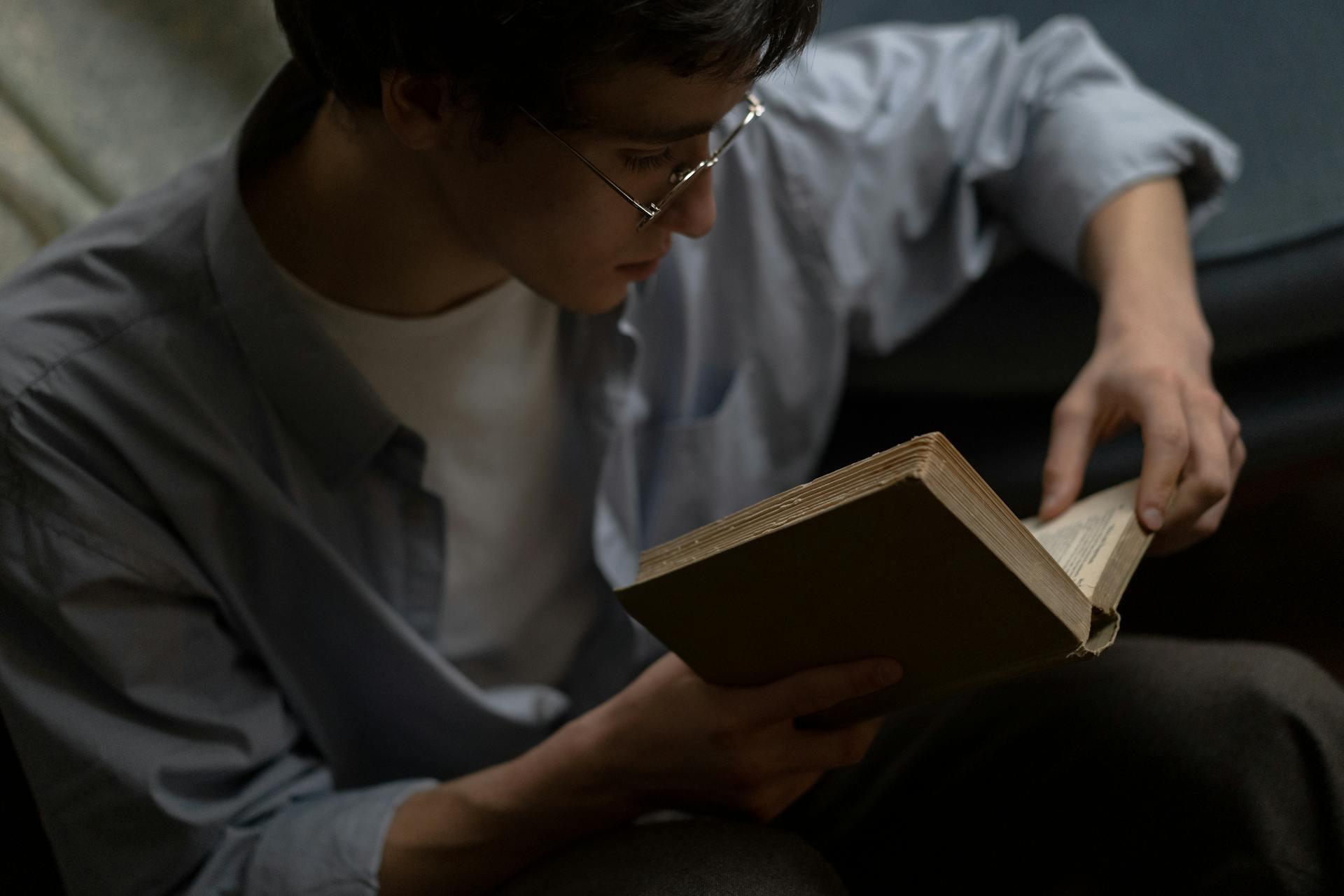 Joven con anteojos y camisa celeste, leyendo un libro.
