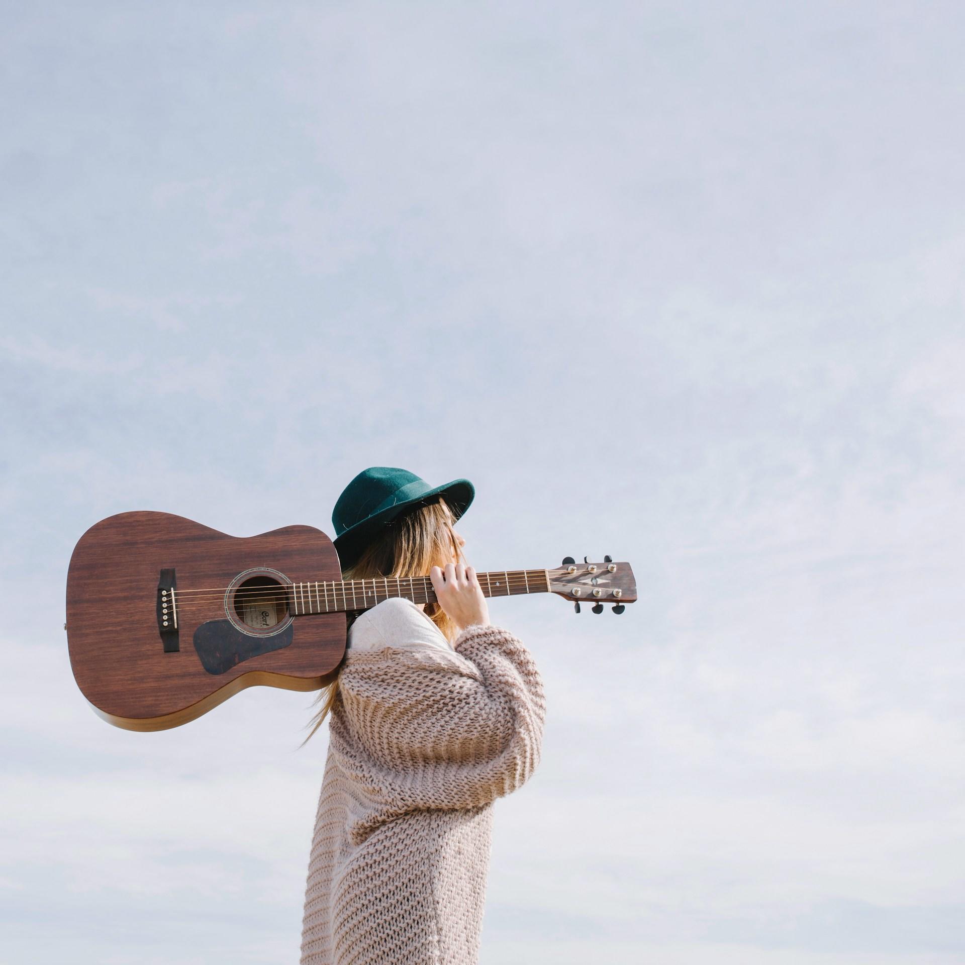 Mujer parada con gorro verde cargando una guitarra sobre su hombro.