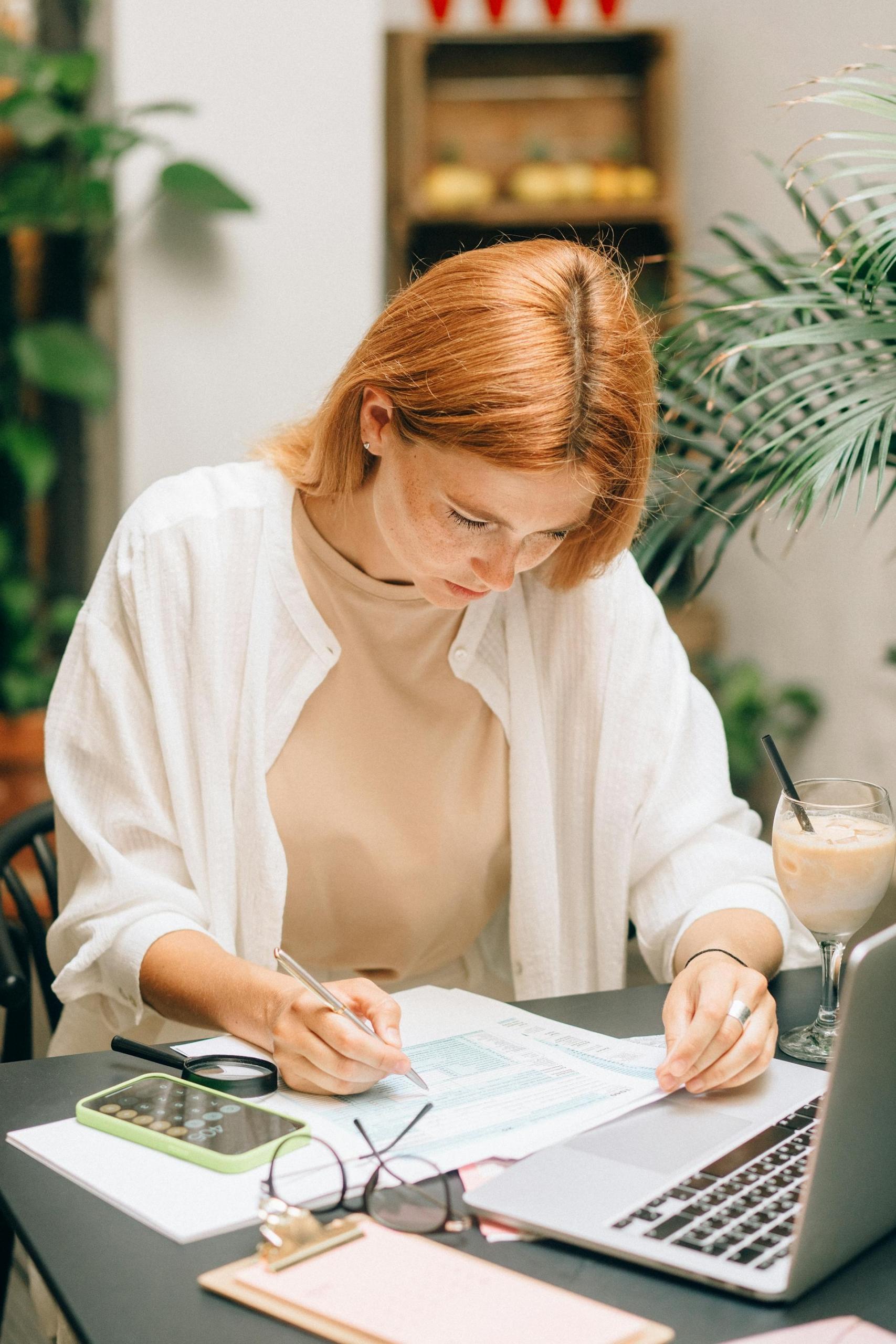 Mujer pelirroja escribiendo en el papel frente a la computadora.