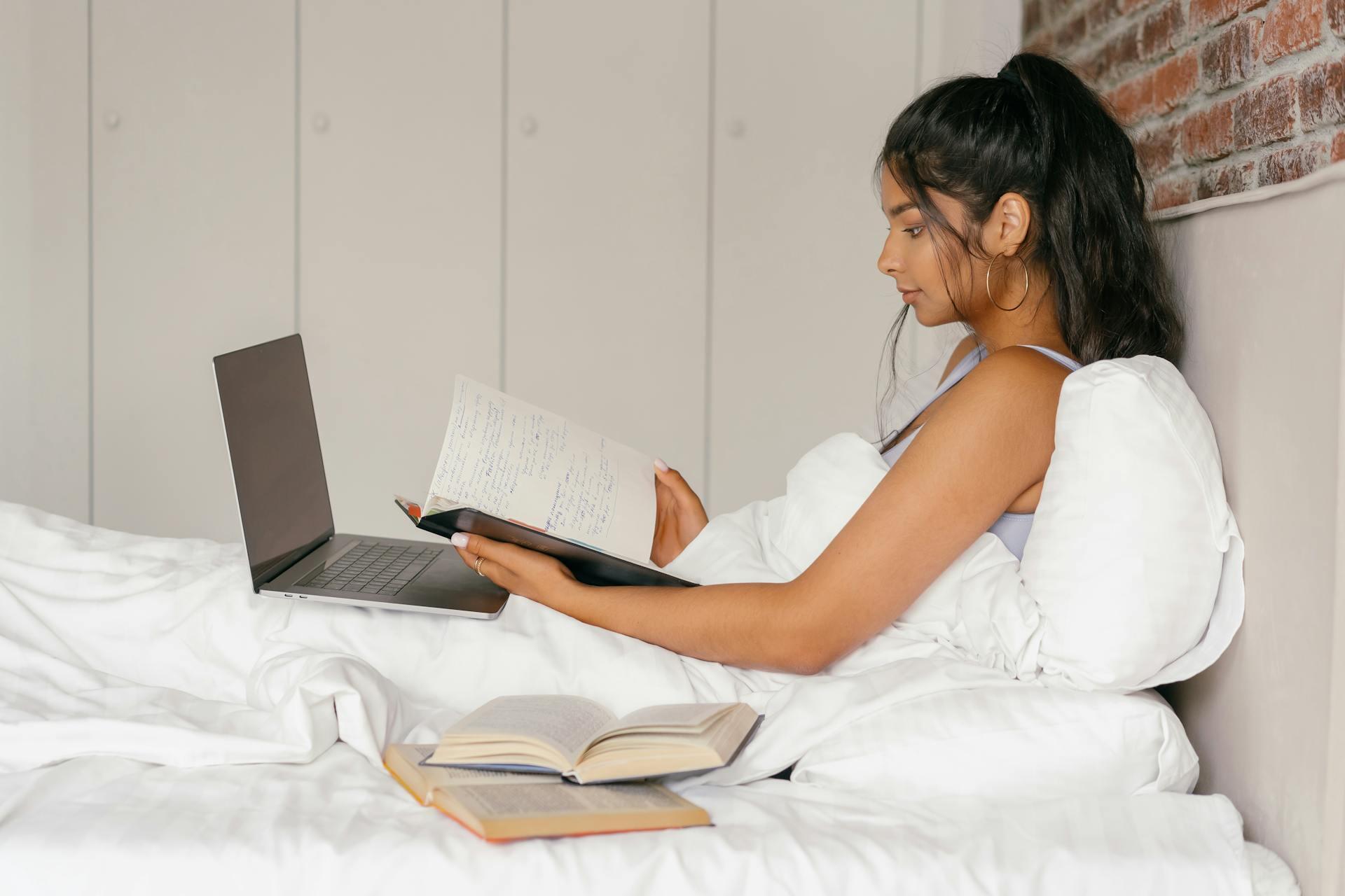 Mujer sentada en la cama con computadora y libros.