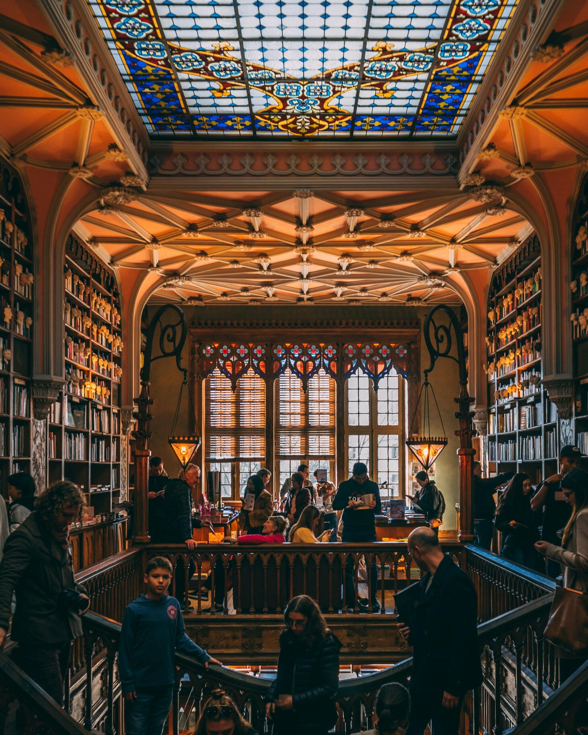 El interior de una gran biblioteca con una arquitectura ornamentada, vidrieras vibrantes y gente mezclándose entre altas estanterías y áreas de lectura.