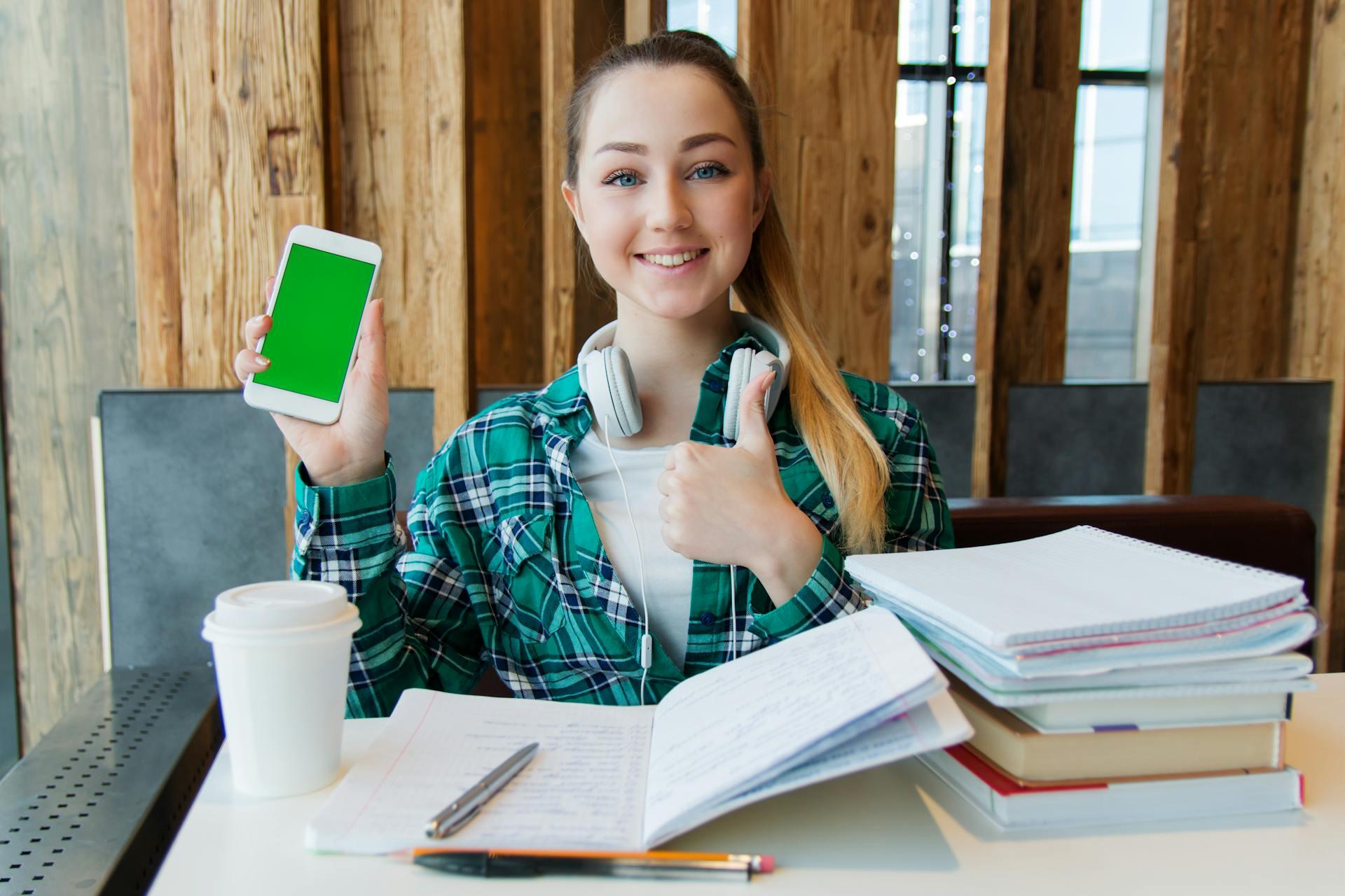 Joven sonriente sentada en el escritorio mostrando el celular.