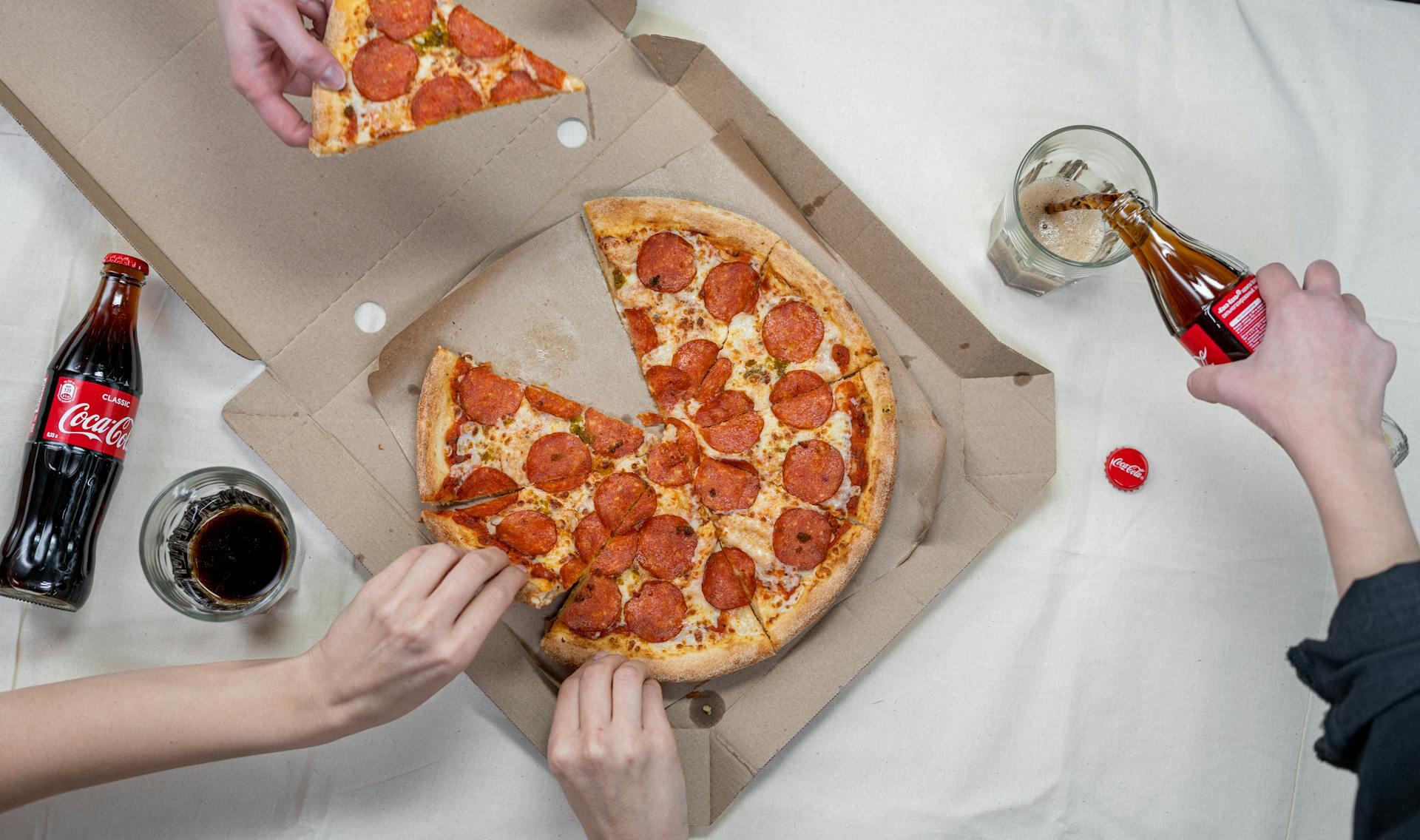 Manos tomando porciones de pizza y sirviendo bebida.