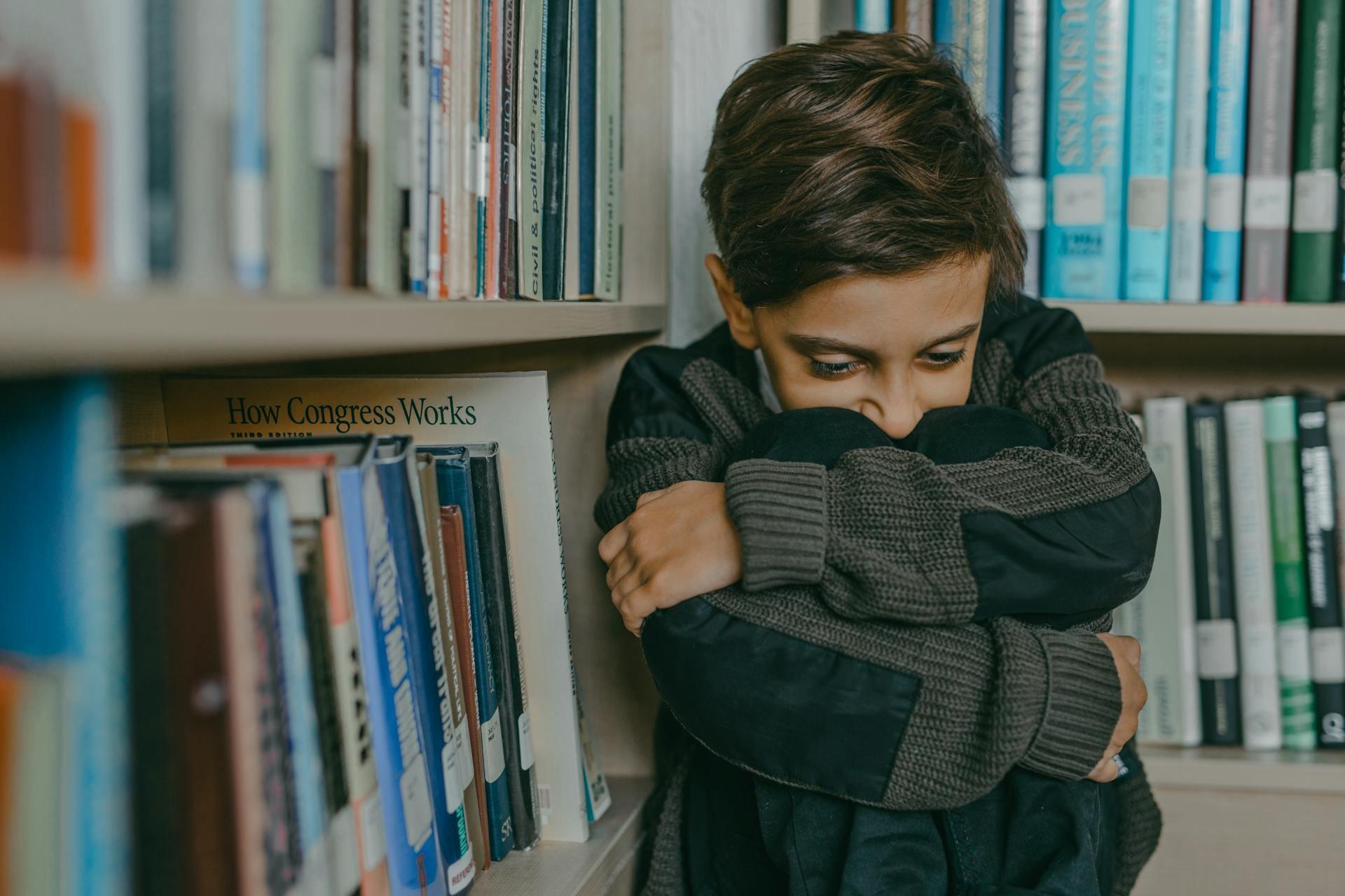 Niño sentado tomando sus piernas con los brazos delante de la biblioteca. 