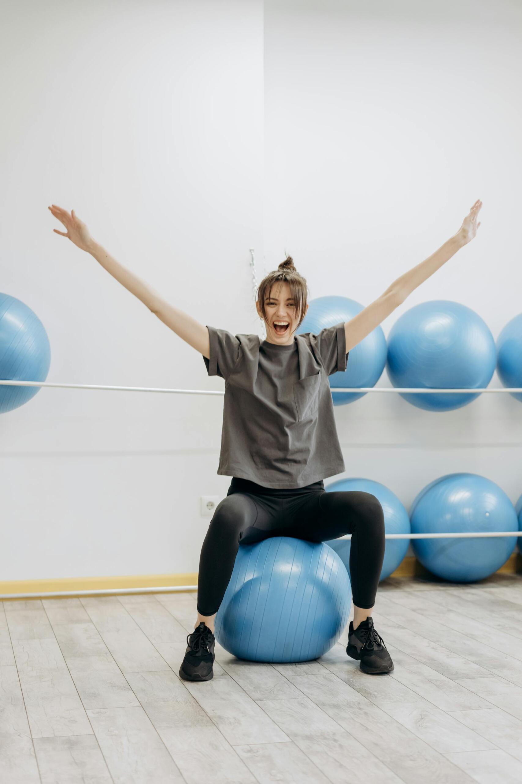 Mujer sonriente sentada sobre una pelota celeste con los brazos levantados.