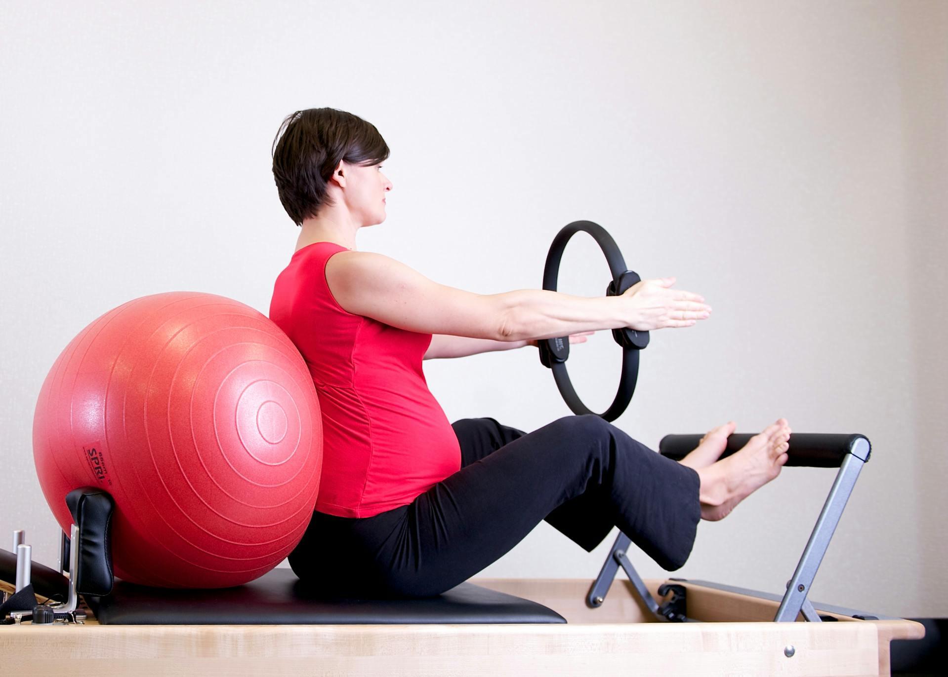 Mujer con remera roja sentada sobre la camilla de pilates apoyando su espalda en una pelota.