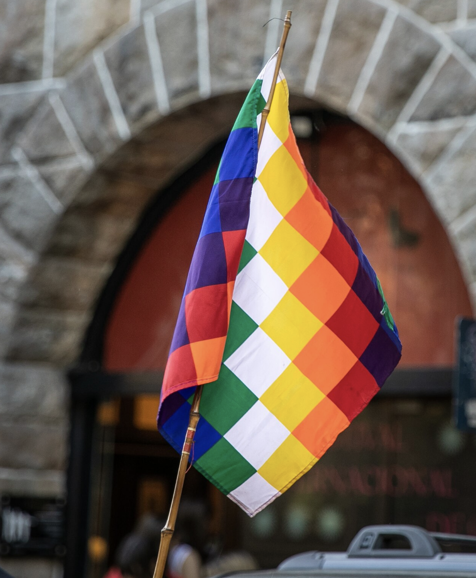 Bandera whipala, símbolo de los pueblos originarios, ondeando frente a un arco de piedra. Fotografía de autor vía Unsplash.