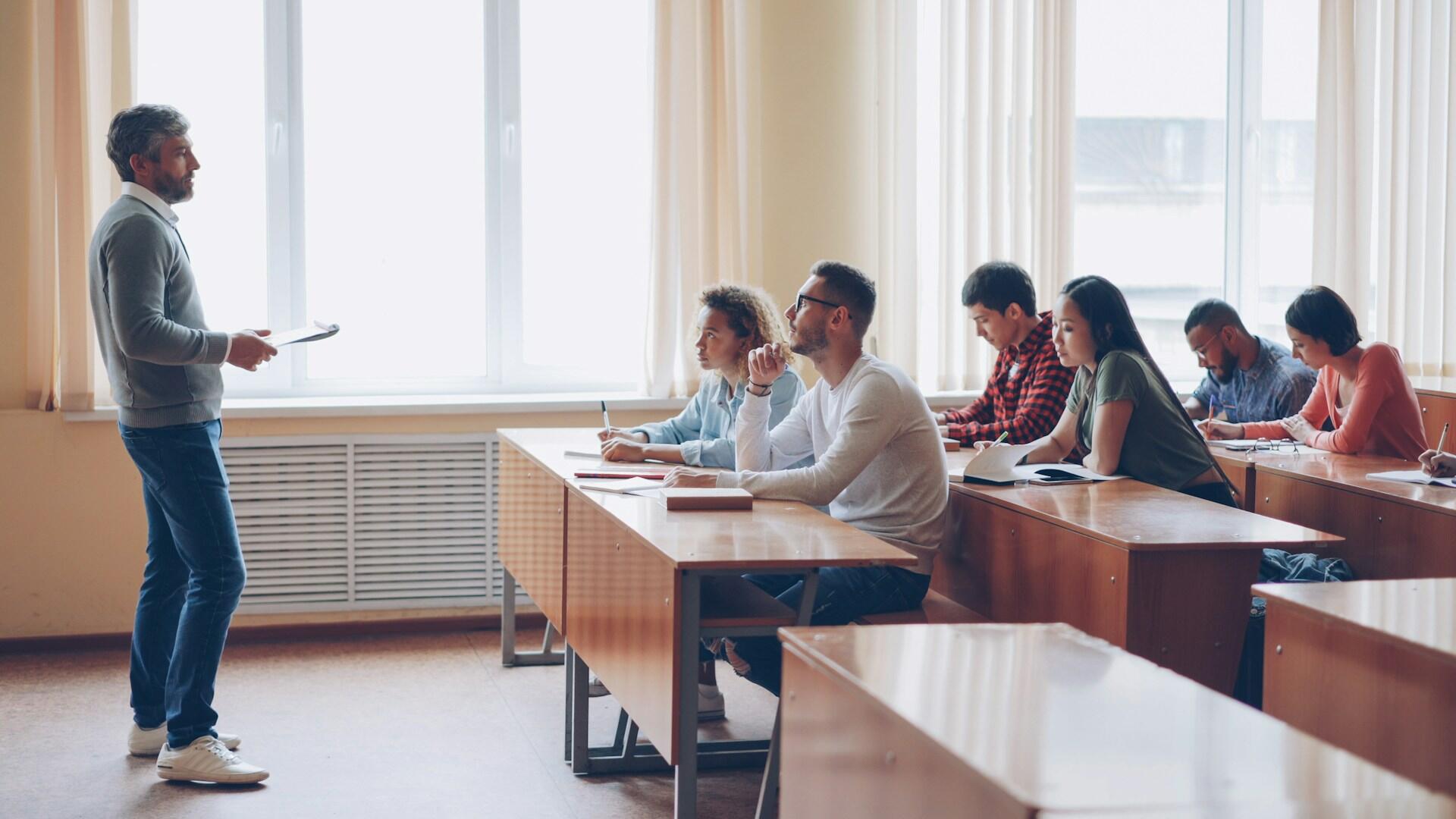 Alumnos sentados en los bancos de la clase escuchando al profesor parado delante.