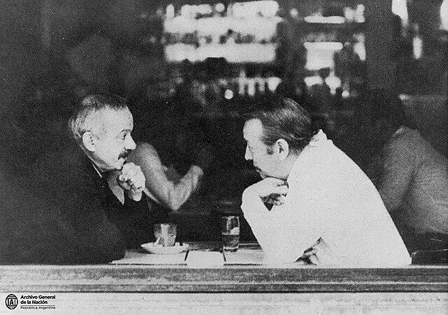 Dos hombres sentados en la mesa detrás del vidrio.