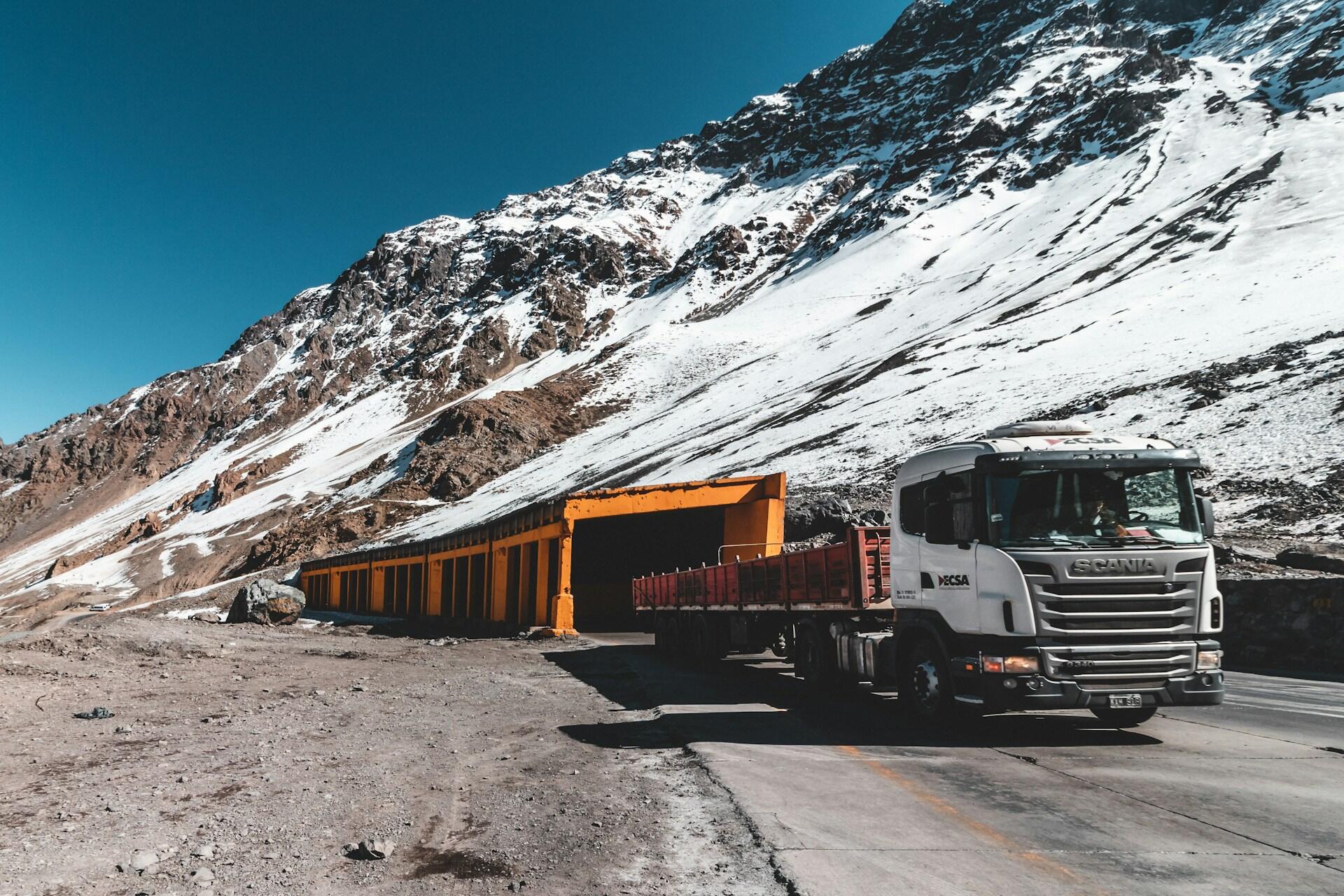 Un camión blanco transita junto a un túnel naranja rodeado de montañas nevadas en los Andes, Chile. Fotografía de Caio Silva.