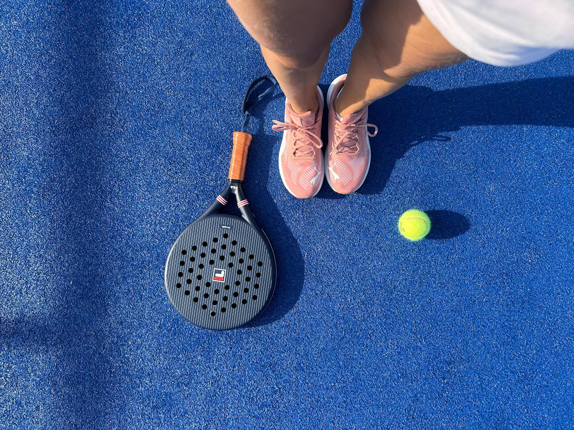 Jugadora de pádel parada sobre una pista azul con una paleta y una pelota de tenis. Fuente: Freepik.