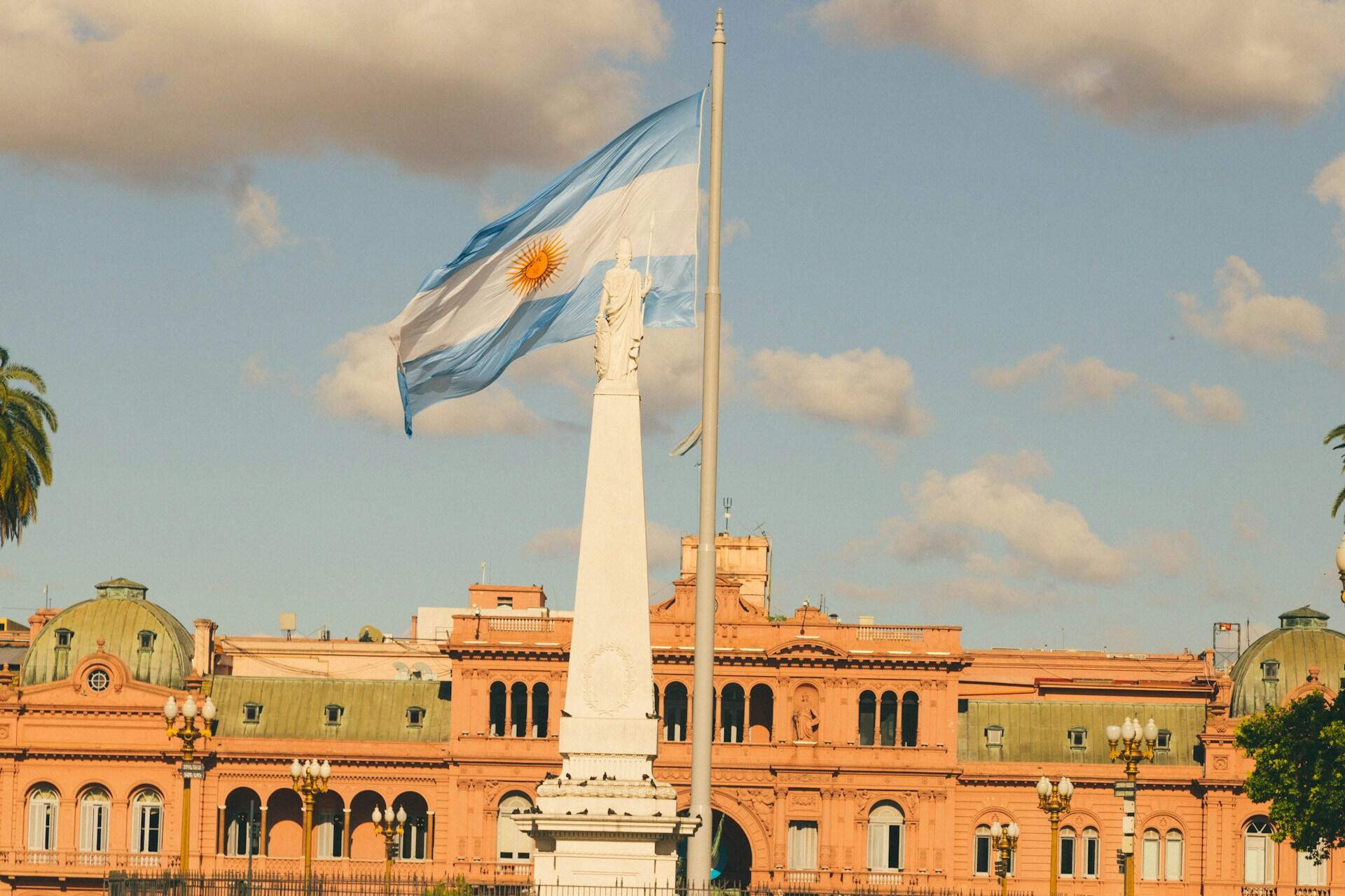 Bandera argentina flameando en la Plaza de Mayo junto a la Pirámide de Mayo y la Casa Rosada en Buenos Aires, representando el centro histórico y político del país. Fotografía de Gustavo Sánchez.