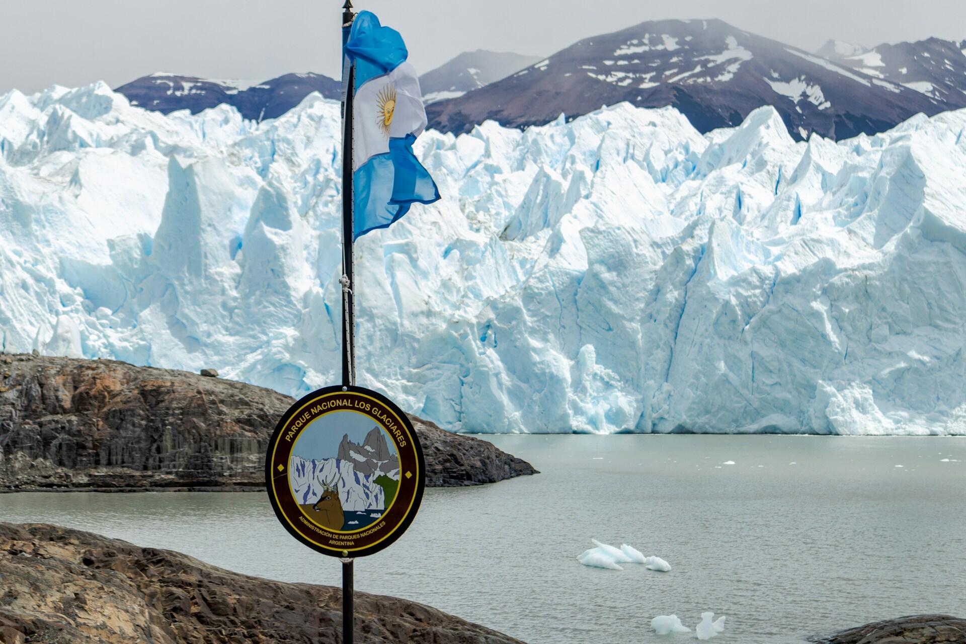 Bandera argentina izada junto al cartel oficial del Parque Nacional Los Glaciares en Santa Cruz con el glaciar de fondo, representando el patrimonio natural nacional. Fotógrafo: Caio Portela.