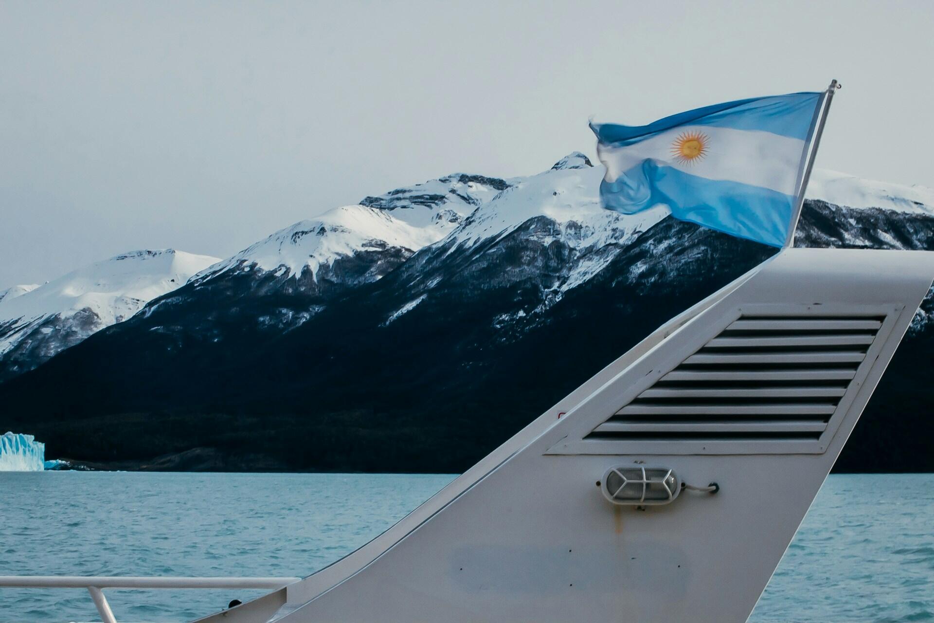 Bandera argentina en la popa de una embarcación navegando frente a montañas nevadas en la Patagonia, representando la soberanía en todo el territorio. Fotógrafo: Felipe Randolfi.