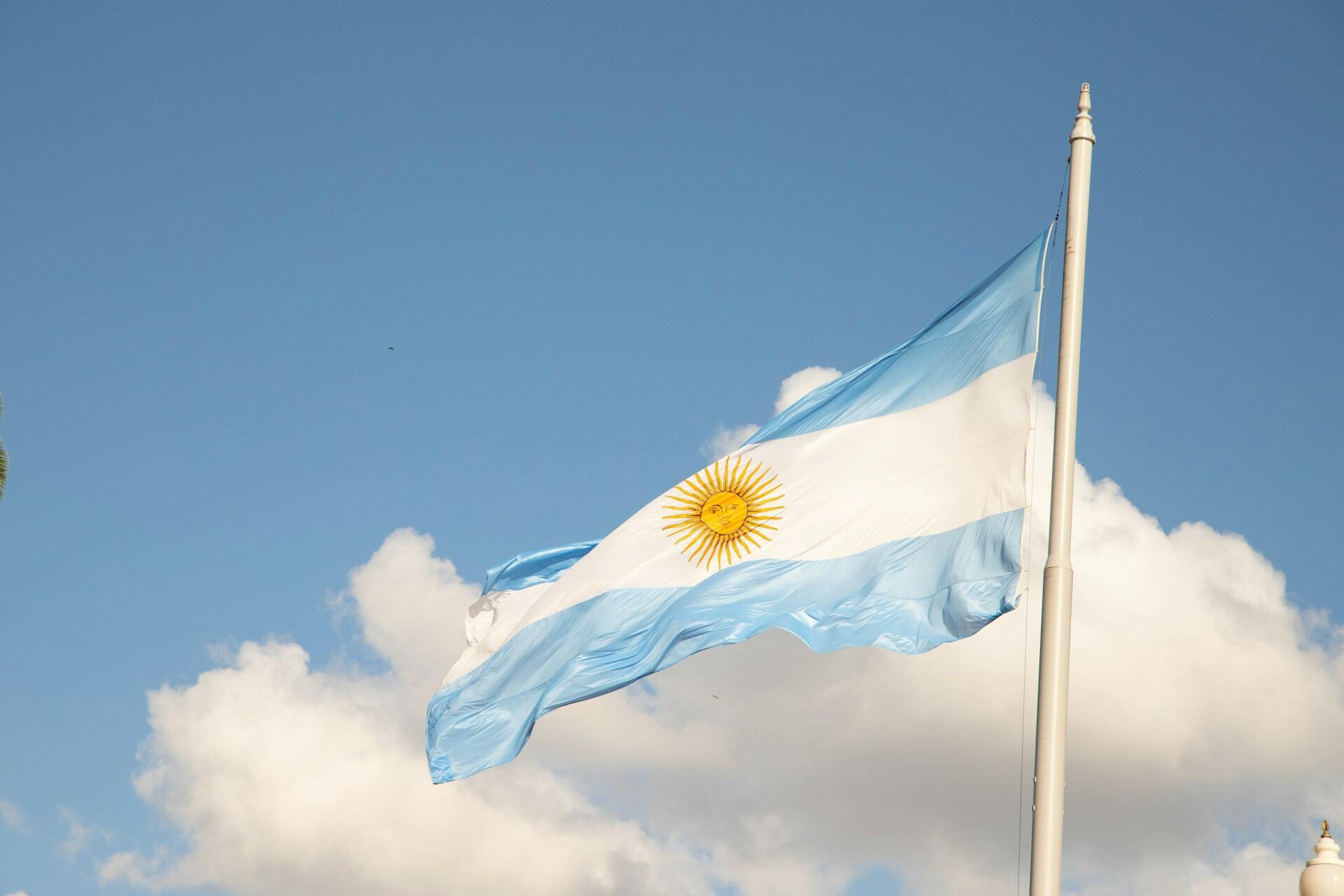 Bandera argentina flameando en lo alto bajo un cielo despejado con nubes en la Ciudad Autónoma de Buenos Aires, representando el orgullo patrio. Fotografía de Gustavo Sánchez.