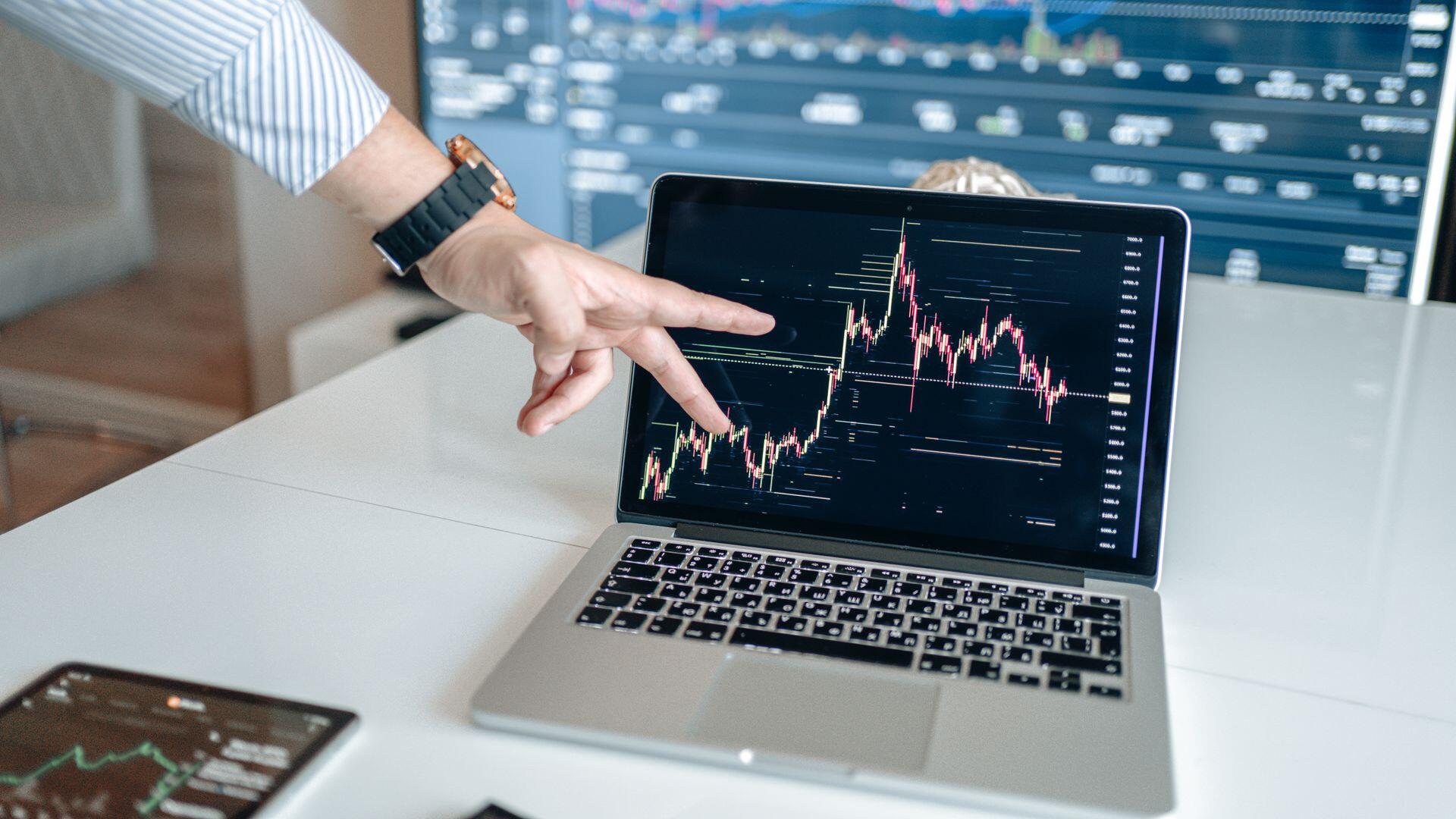 Oficina de inversiones, mano señalando gráficos de velas financieras en una laptop, AlphaTradeZone.