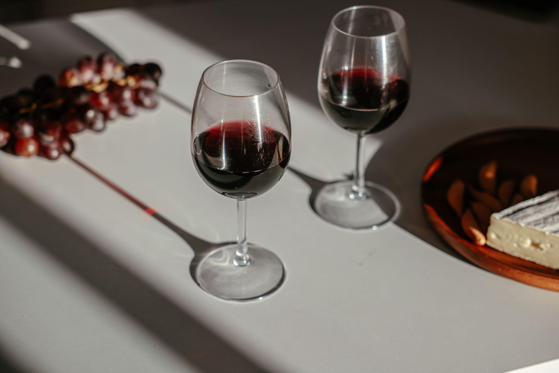 Dos copas de vino tinto y un racimo de uvas apoyados en la superficie blanca.
