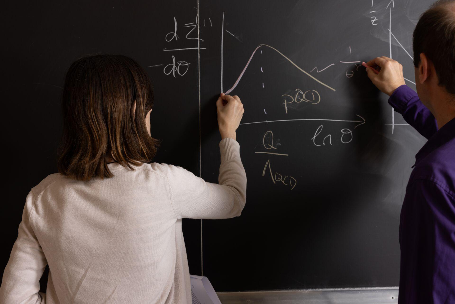 Aula de estudio con dos personas desarrollando gráficos y fórmulas complejas en un pizarrón negro. Fuente: Pexels.