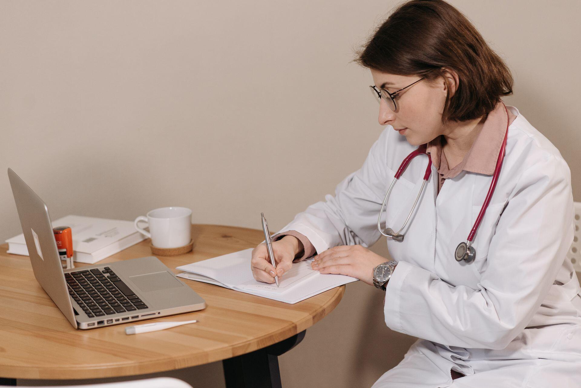 Escritorio médico, profesional de la salud redactando un informe junto a su computadora, por Pavel Danilyuk.