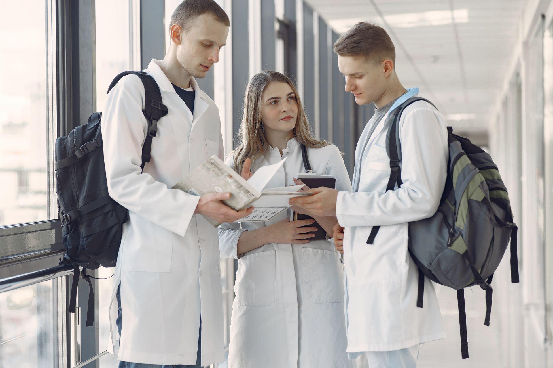 Pasillo de facultad de medicina, tres estudiantes con batas blancas consultando un libro de texto, Gustavo Fring.
