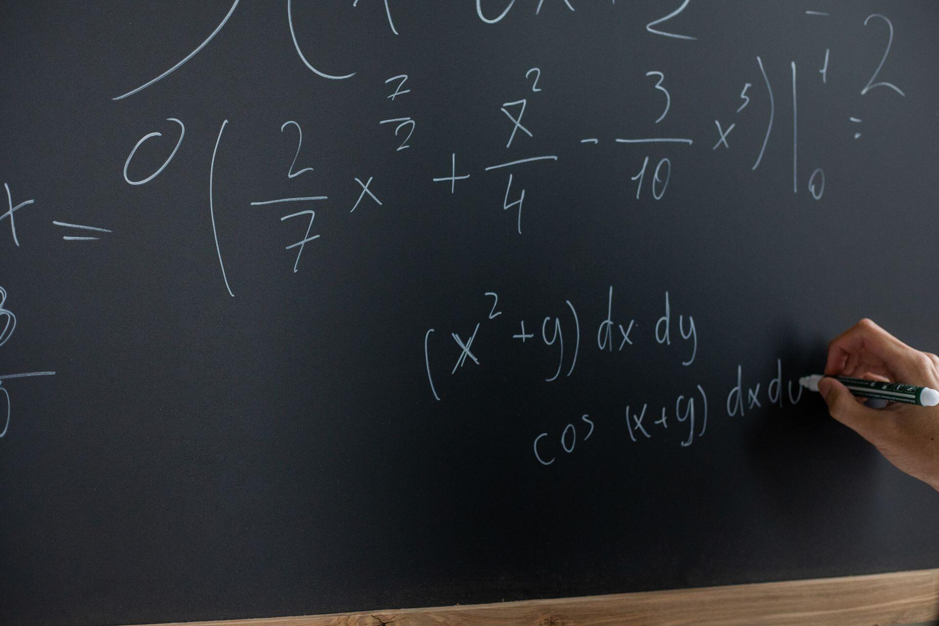 Primer plano de una mano escribiendo ecuaciones y derivadas con tiza sobre un pizarrón oscuro. Fuente: Pexels.