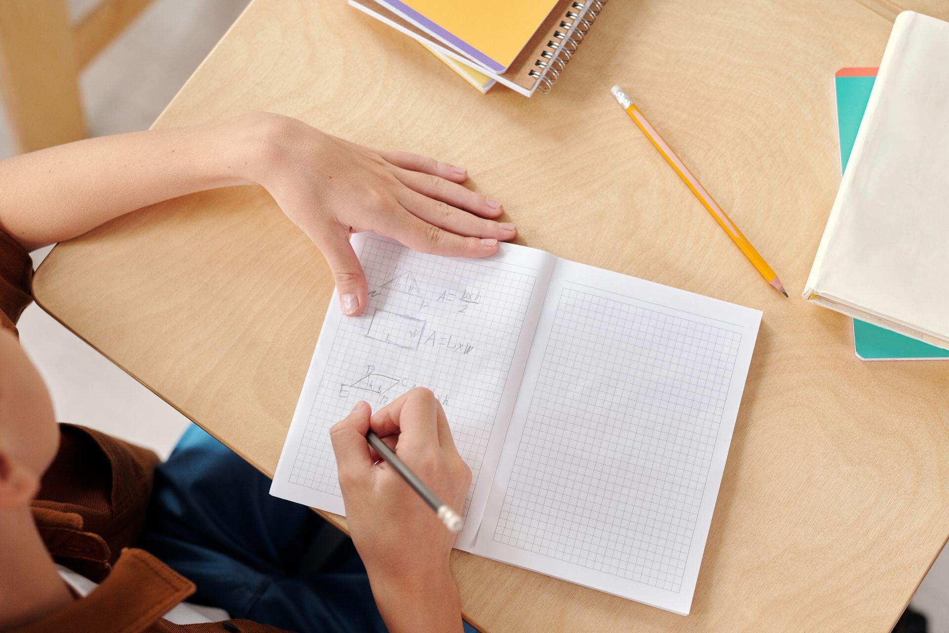 Plano cenital de un escritorio donde una mano dibuja figuras geométricas en un cuaderno cuadriculado. Fuente: Pexels.